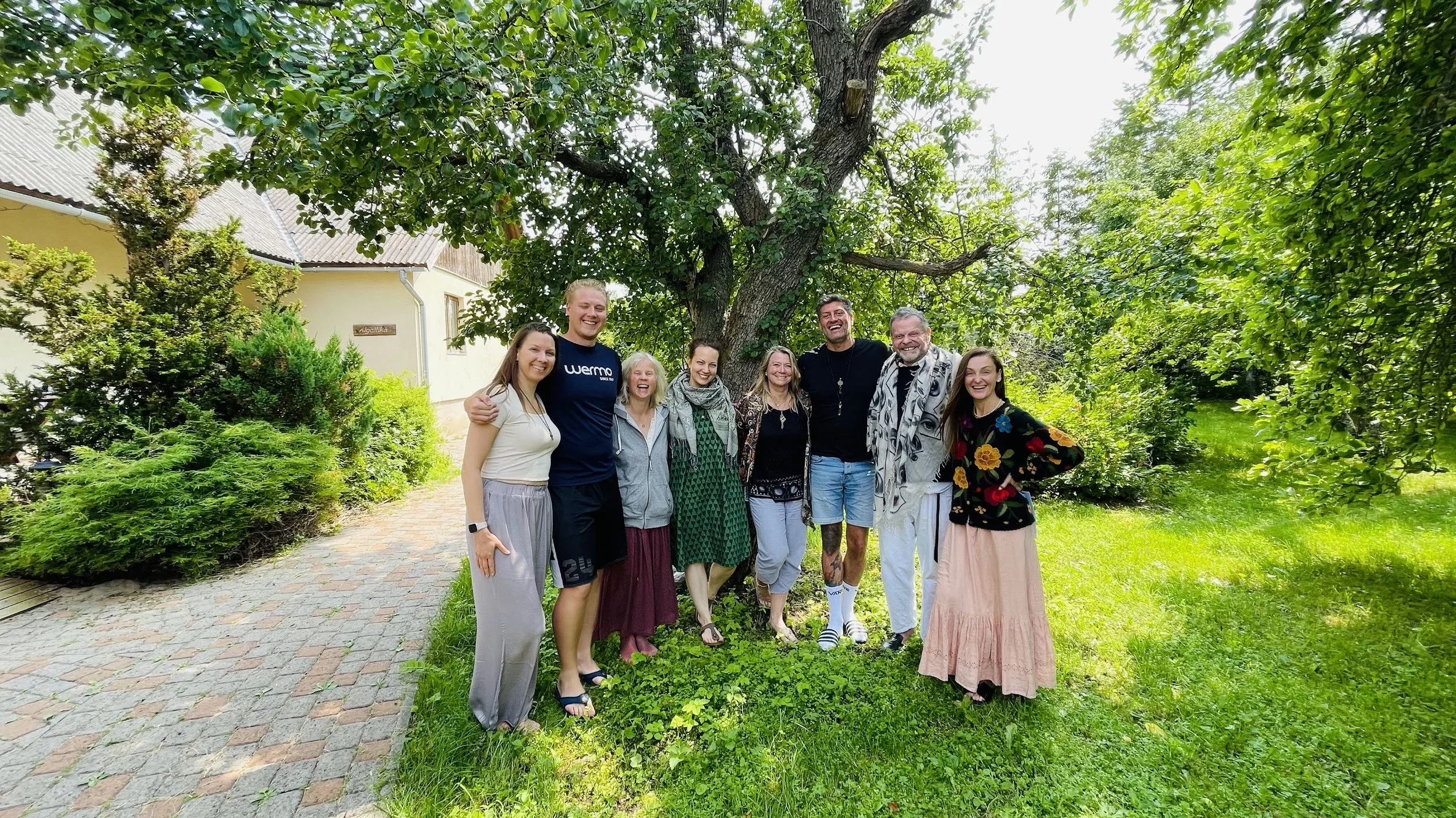 Group of nine diverse people standing outdoors under a large leafy tree, smiling and posing for a photo on a sunny day.