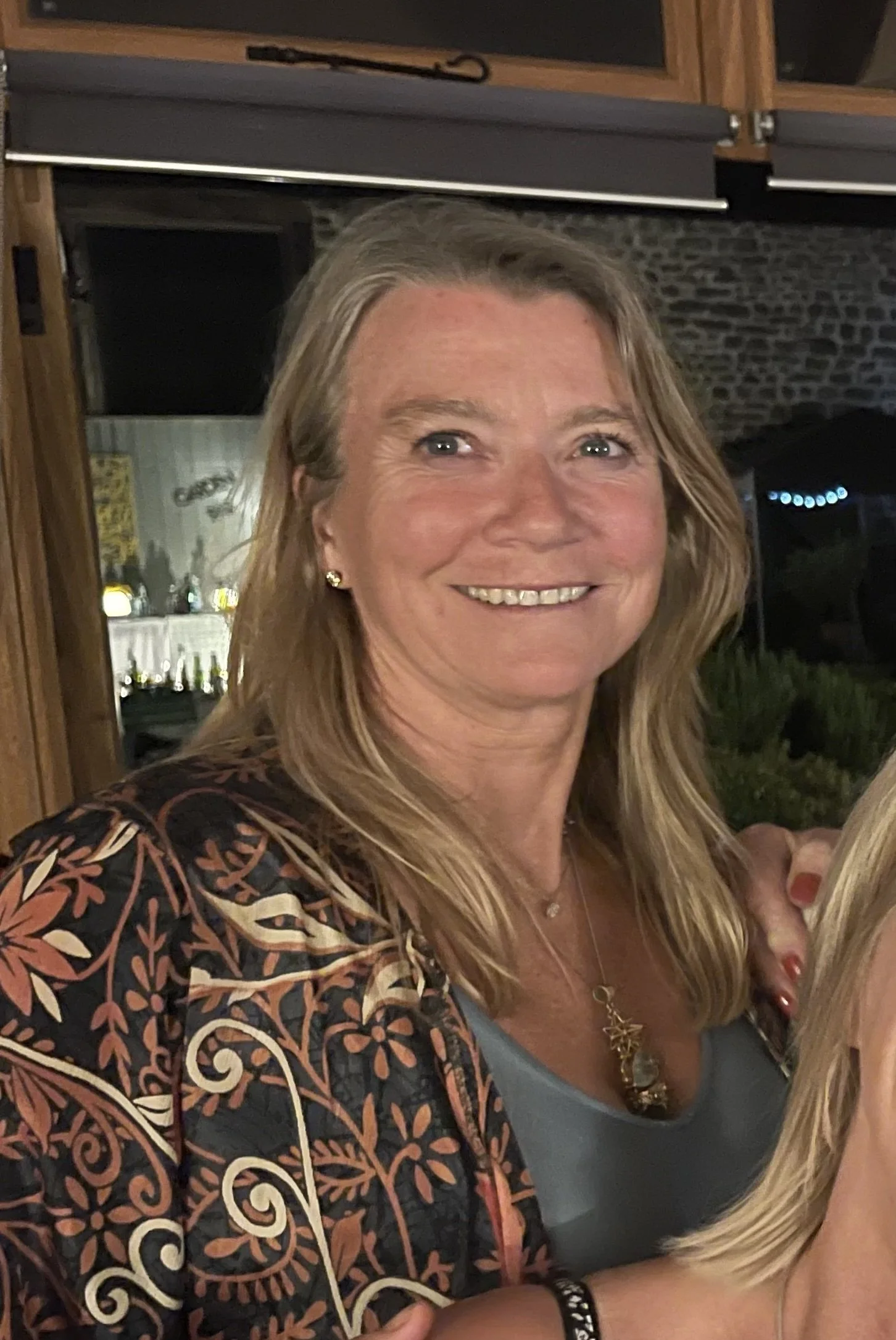 A smiling woman with shoulder-length blonde hair, wearing a patterned jacket over a gray top, standing indoors at a social gathering.