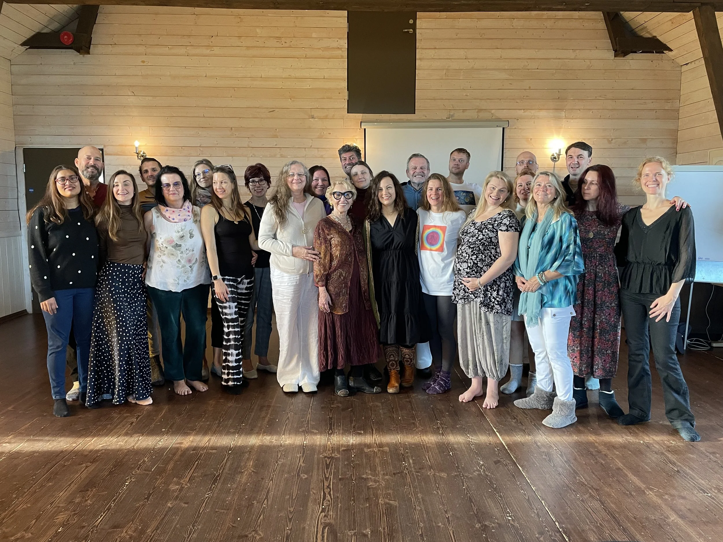 Group of people standing on a wooden floor in a room with wooden walls, smiling for a photo.