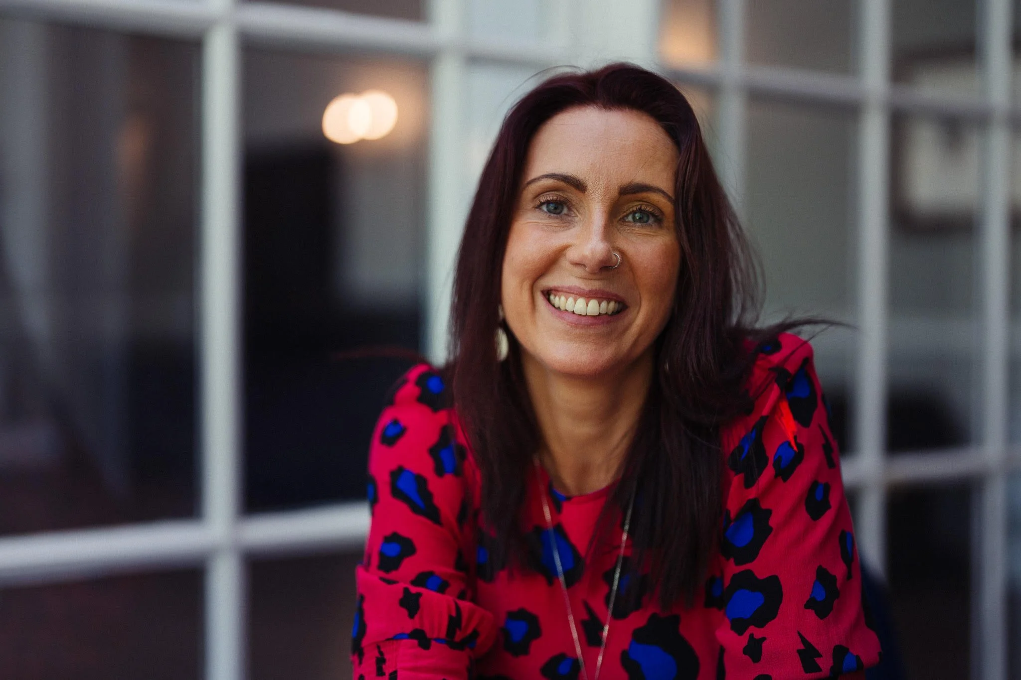 A woman with reddish-brown hair, smiling, wearing a pink blouse with blue and black leopard print, sitting indoors near a glass wall or window.