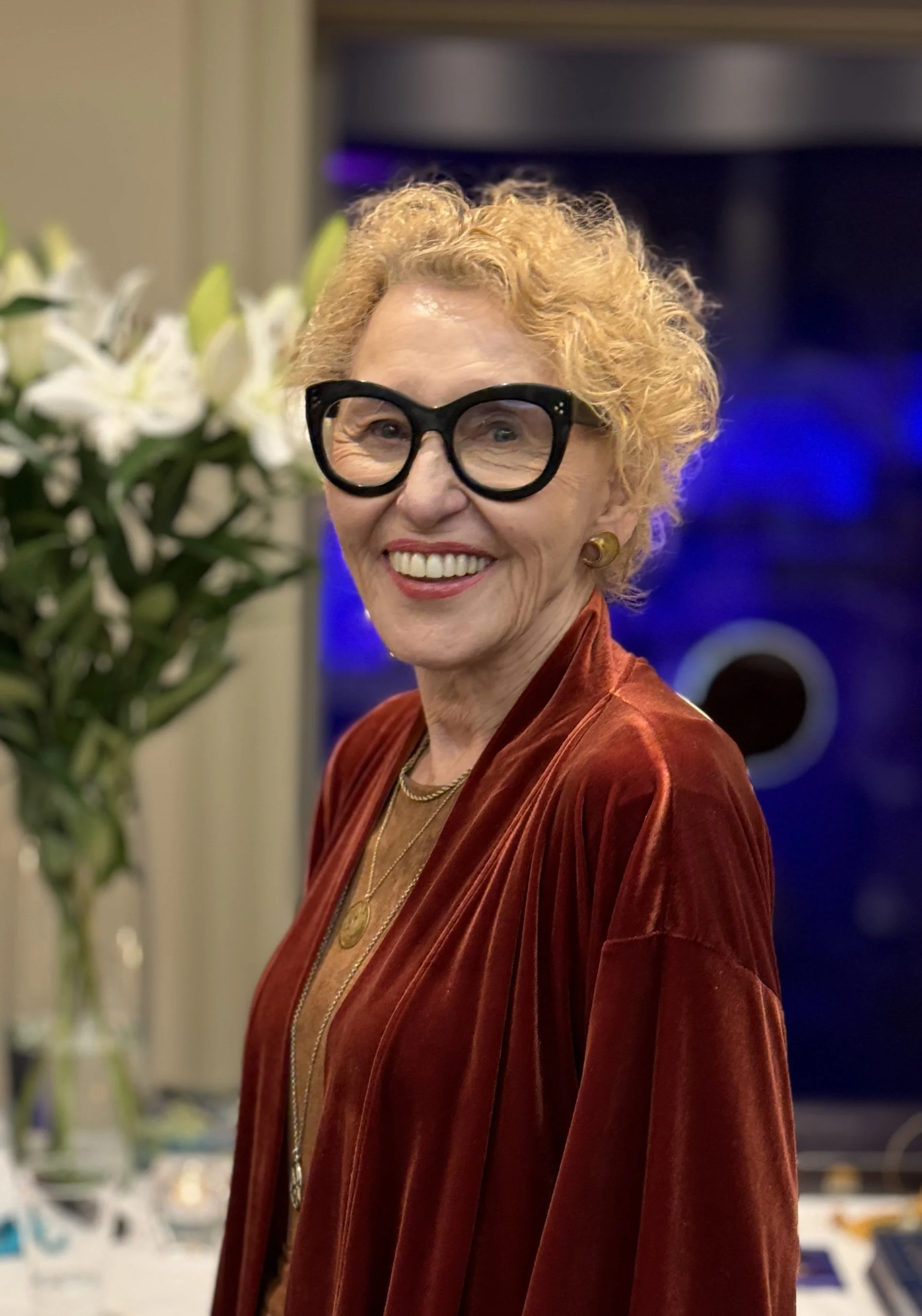 An elderly woman with curly blonde hair, wearing large black glasses, a red velvet jacket, and gold jewelry, smiling at a social event with white lilies in the background.