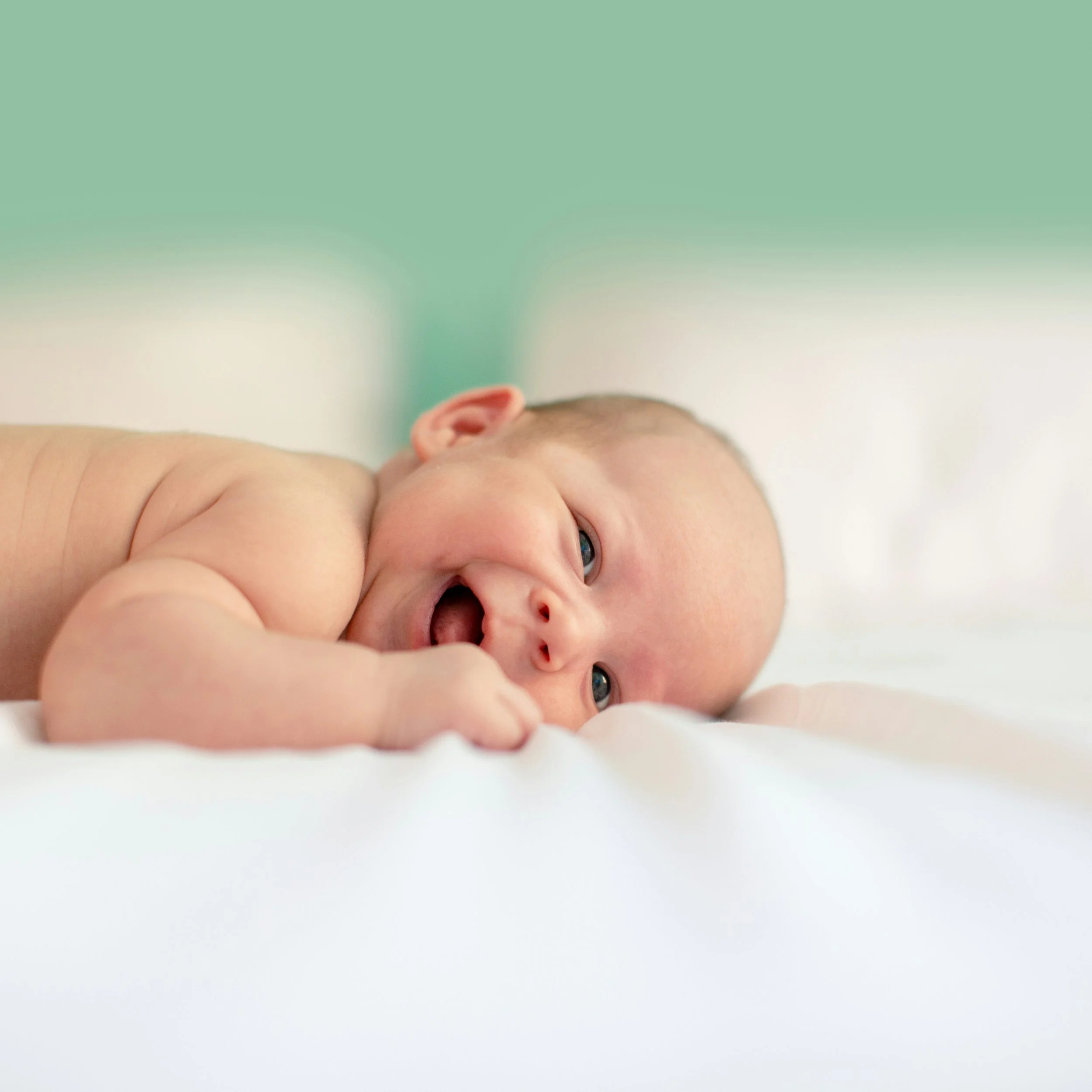 Close-up of a newborn baby lying on a white surface, looking at the camera with bright eyes and a slightly open mouth