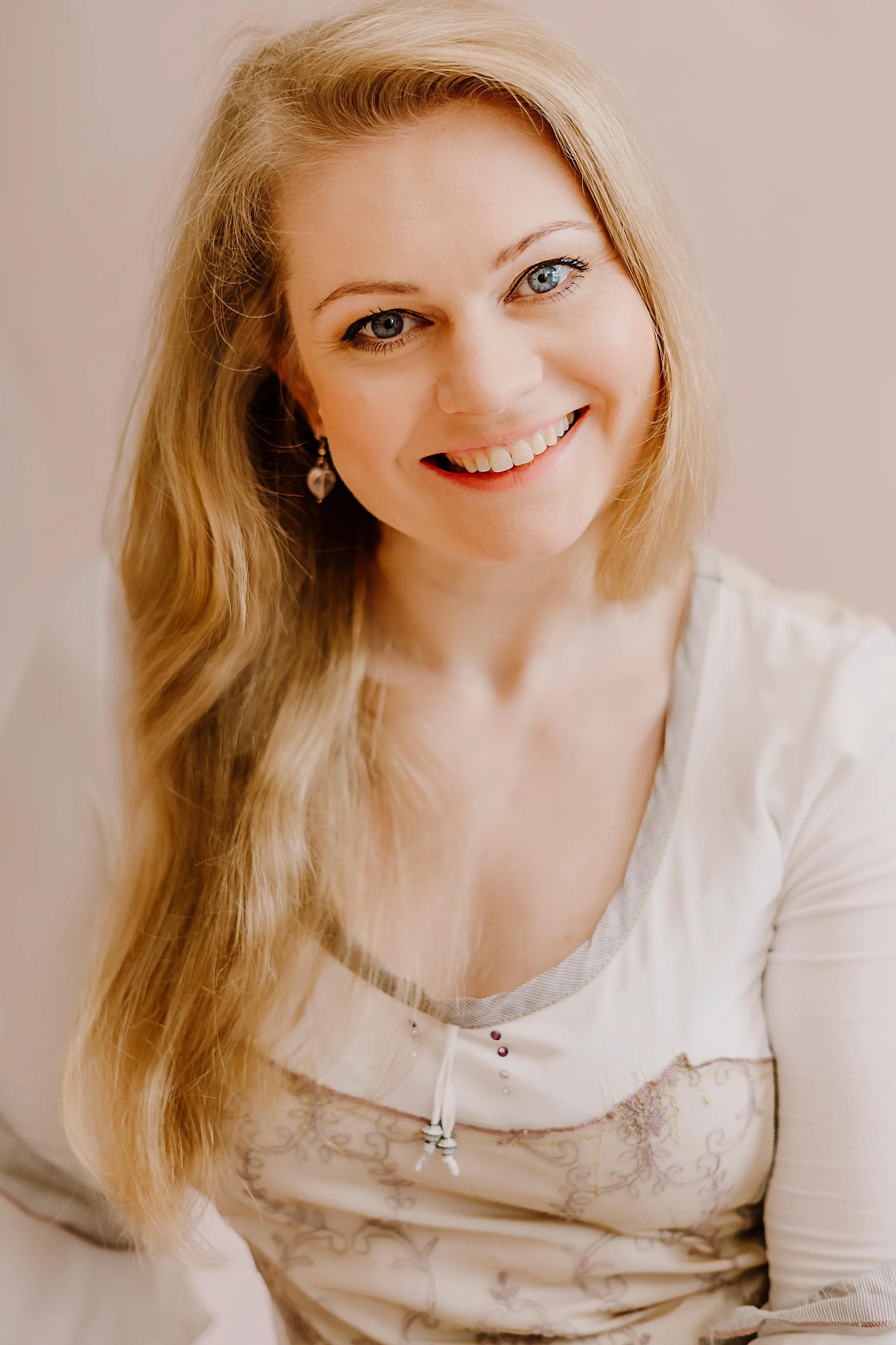 A smiling woman with long blonde hair, blue eyes, and earrings, wearing a cream-colored shirt with decorative details.