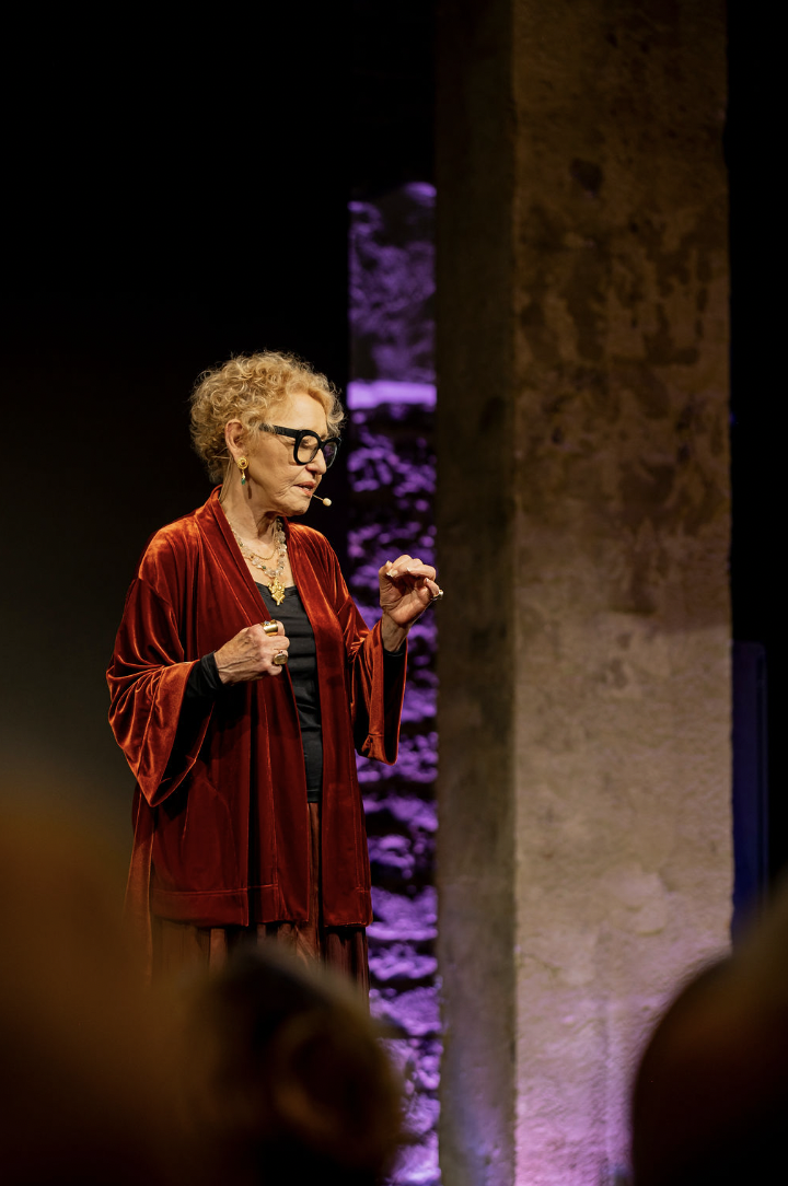 Older woman with curly blonde hair wearing glasses and a headset microphone, dressed in a red velvet jacket and jewelry, speaking on a stage with purple and beige lighting.