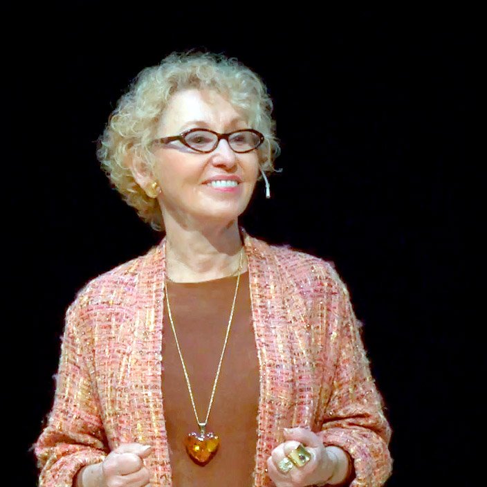 A smiling woman with curly blonde hair, wearing glasses, a beige blazer over a brown top, and holding her hands in front of her. She is accessorized with a long gold chain with a heart-shaped pendant, earrings, and rings. The background is black.