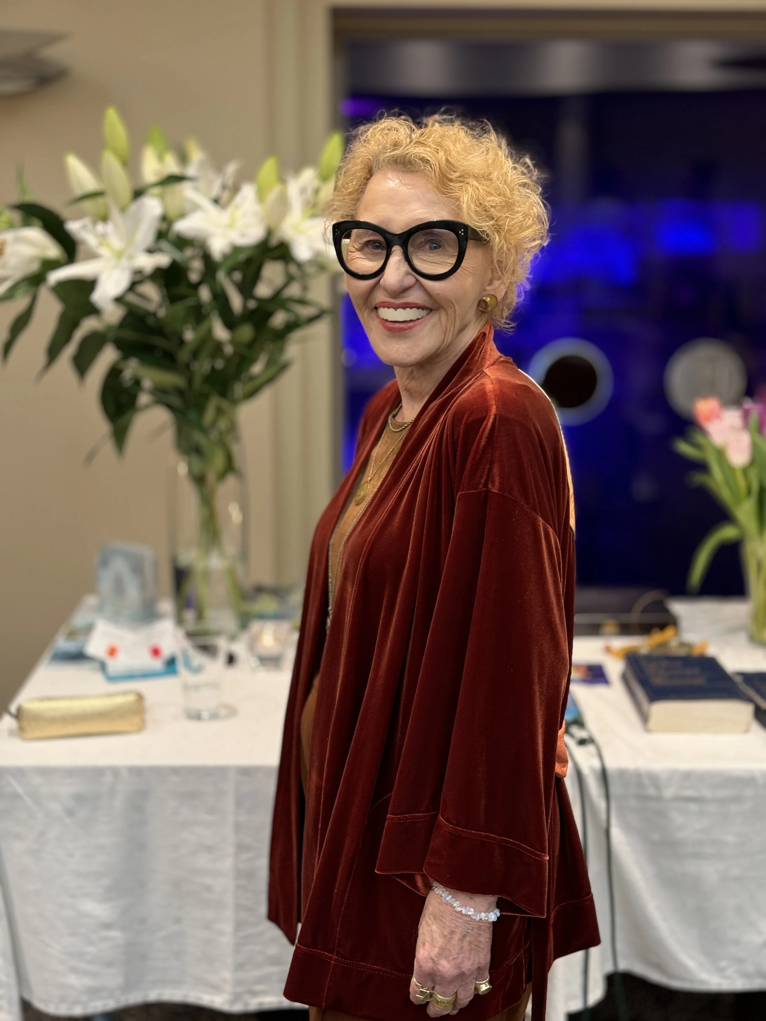 An elderly woman with short, curly blonde hair and large black glasses standing indoors with a smile, wearing a rust-colored velvet jacket and jewelry, with a table of flowers and books in the background.