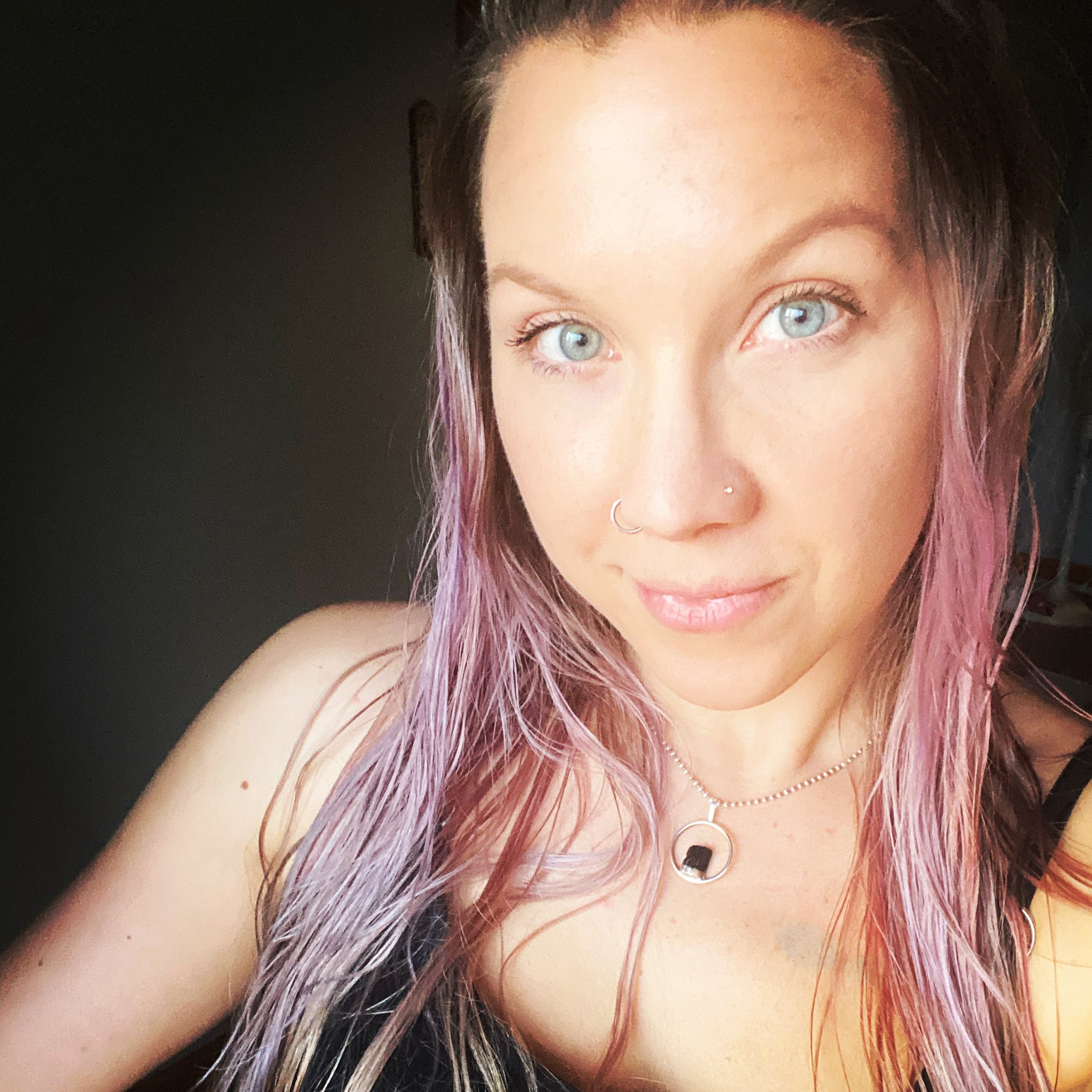 A woman with long, pink-tinted hair, blue eyes, and a nose piercing, smiling inside near a window with sunlight on her face.