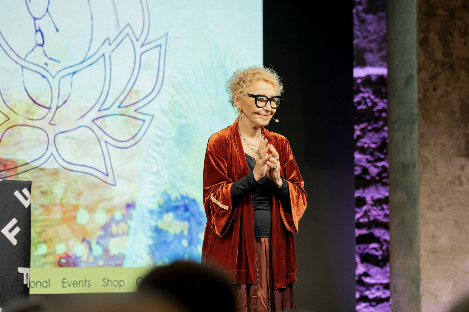 An elderly woman with short curly blonde hair, wearing large black glasses and a red velvet jacket, stands on stage with her hands clasped near her chest, smiling gently while speaking into a microphone headset. Behind her is a colorful large screen with a leaf design.