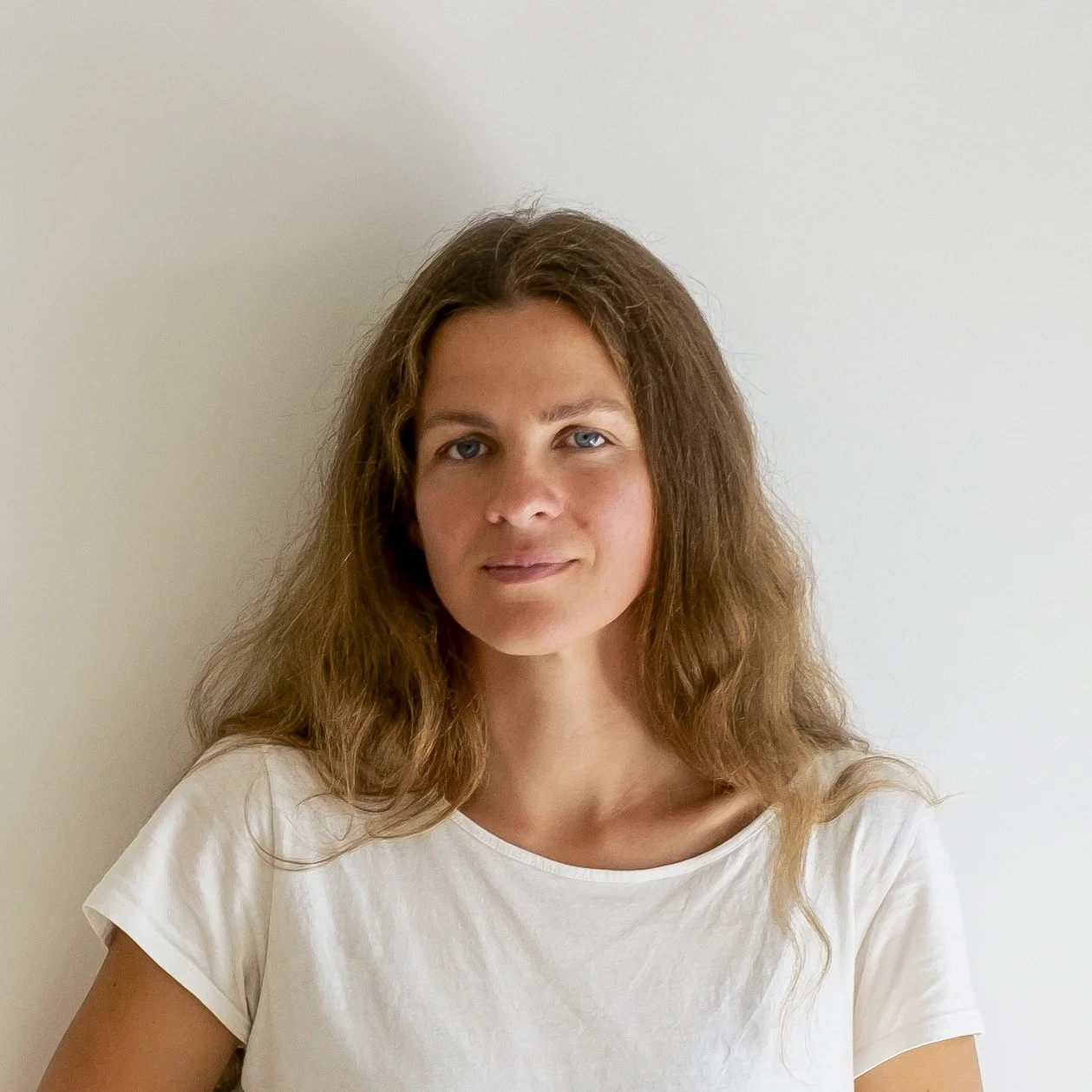 A woman with long, wavy brown hair, blue eyes, and light skin, wearing a white t-shirt, standing against a plain white wall.