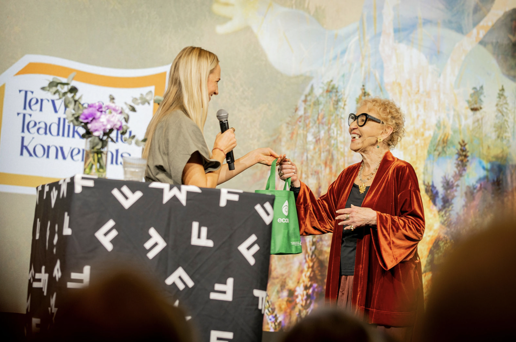 Two women, one younger and one older, exchanging a gift on stage at a conference, with a backdrop displaying 'Terry Teadlink Konvergens'. The younger woman is smiling and holding a microphone, while the older woman is dressed in a red velvet jacket and wearing glasses.