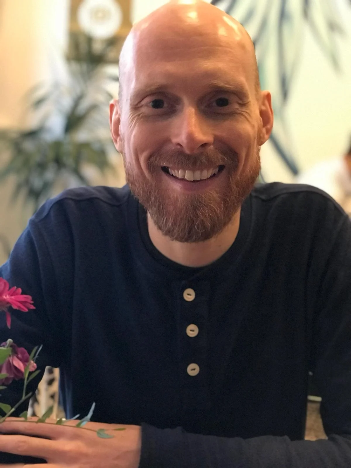 A smiling man with a bald head and a beard, sitting at a table with pink flowers. He is wearing a navy blue shirt with white buttons.