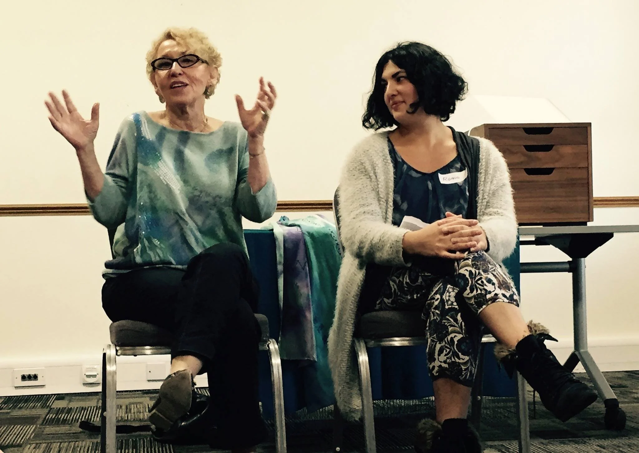 Two women sitting on chairs engaged in conversation; one woman with curly blonde hair and glasses gesturing with her hands, the other woman with curly black hair and a name tag, listening attentively.