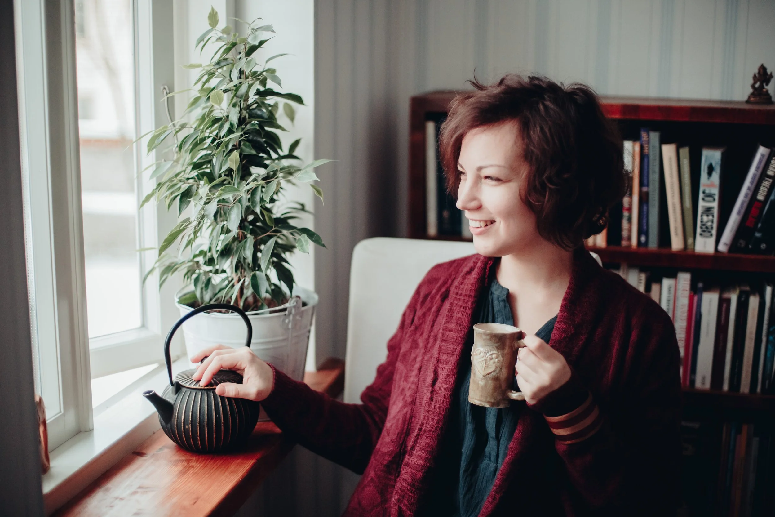 A woman with short, curly brown hair smiling while sitting by a window, holding a ceramic mug in her right hand, with a teapot on the windowsill and a large leafy plant nearby.