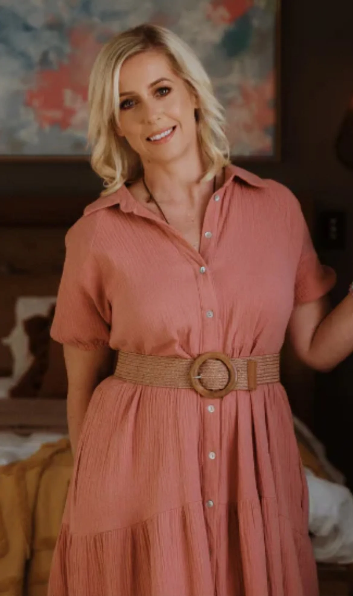 A woman with shoulder-length blonde hair smiling, wearing a peach-colored dress with a matching belt, standing inside a room with a colorful abstract painting on the wall behind her.