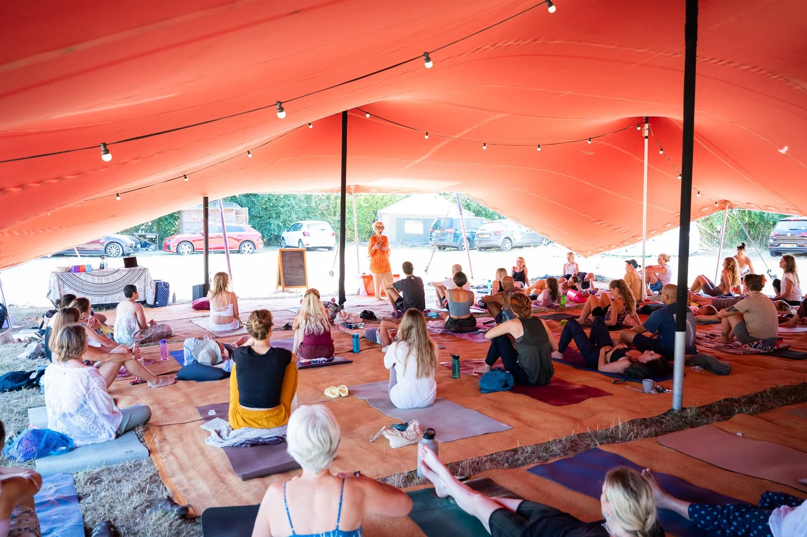 People participating in a yoga class under a large orange tent outdoor, sitting on yoga mats and listening to an instructor at the front. There are parked cars and green trees outside the tent.