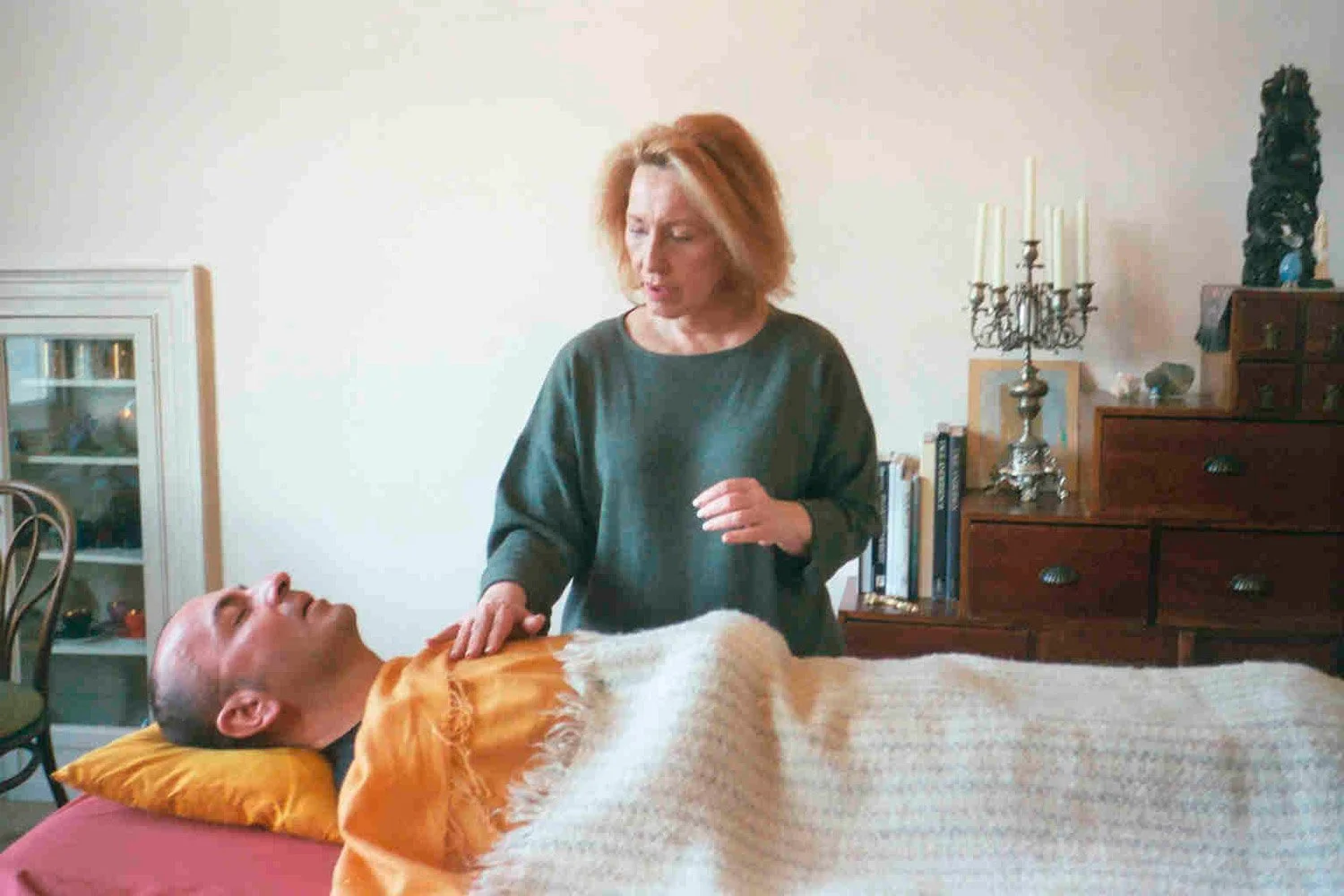 A woman stands next to a man lying in bed, touching his chest gently. The man appears to be unresponsive, and the scene looks like a caregiving moment in a bedroom.