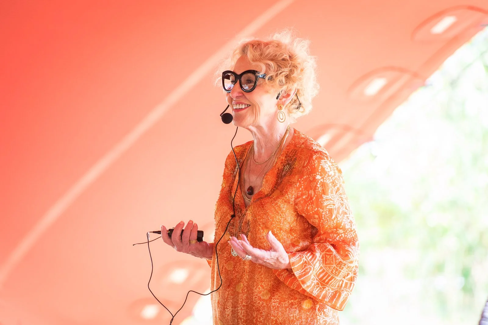 A smiling elderly woman with curly blonde hair, wearing glasses and an orange patterned blouse, giving a presentation using a headset microphone.