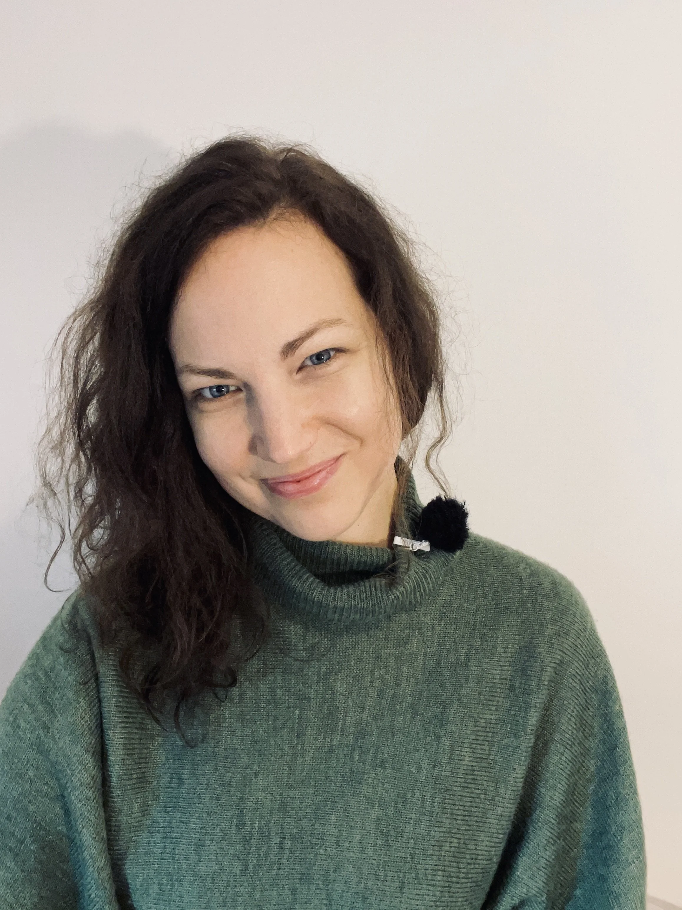 A woman with wavy brown hair and blue eyes, wearing a green turtleneck sweater and smiling, standing against a plain white background.