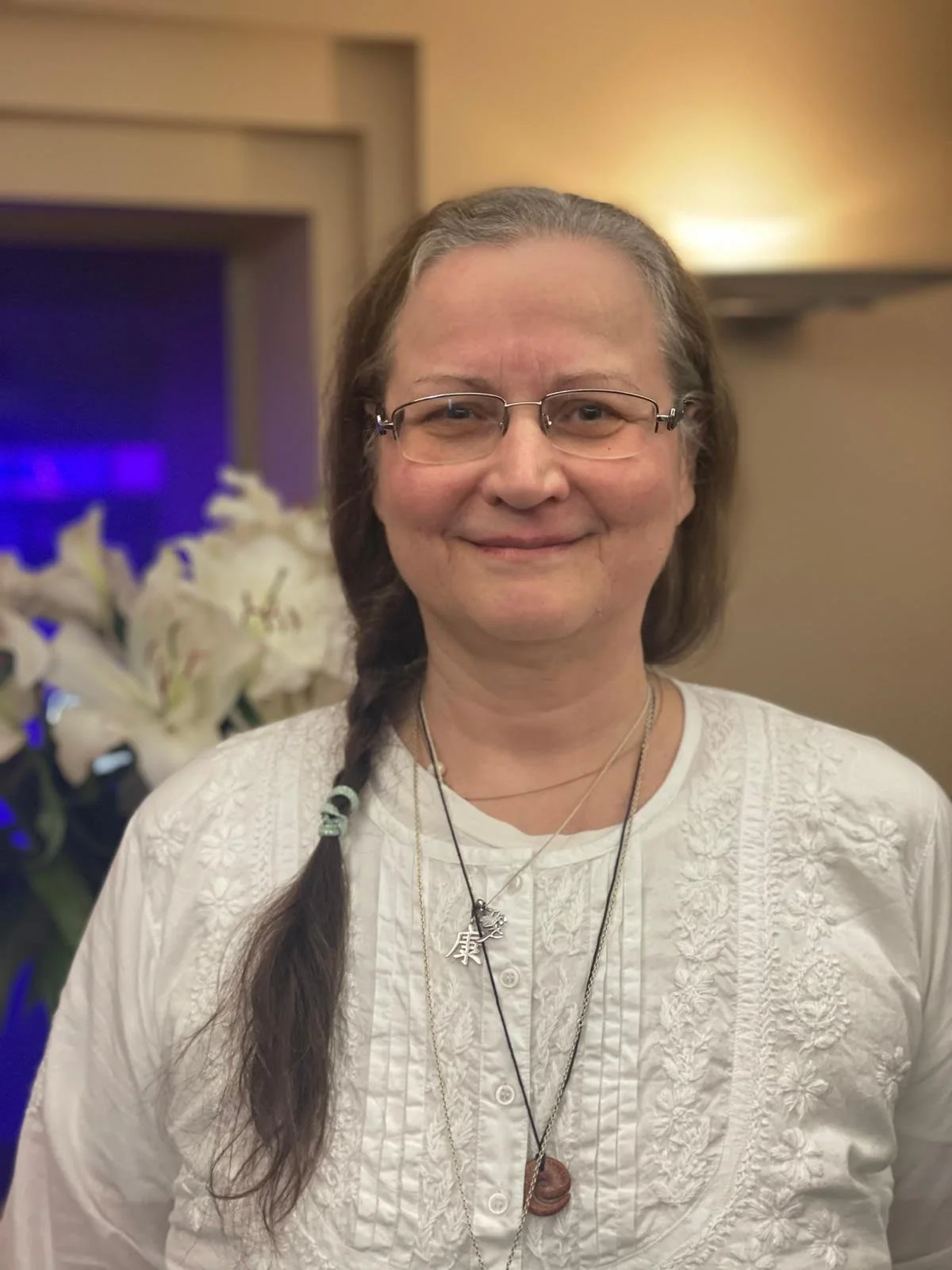 A smiling middle-aged woman with glasses and long brown hair in a braid, wearing a white embroidered blouse with layered necklaces, standing indoors near a bouquet of white lilies.