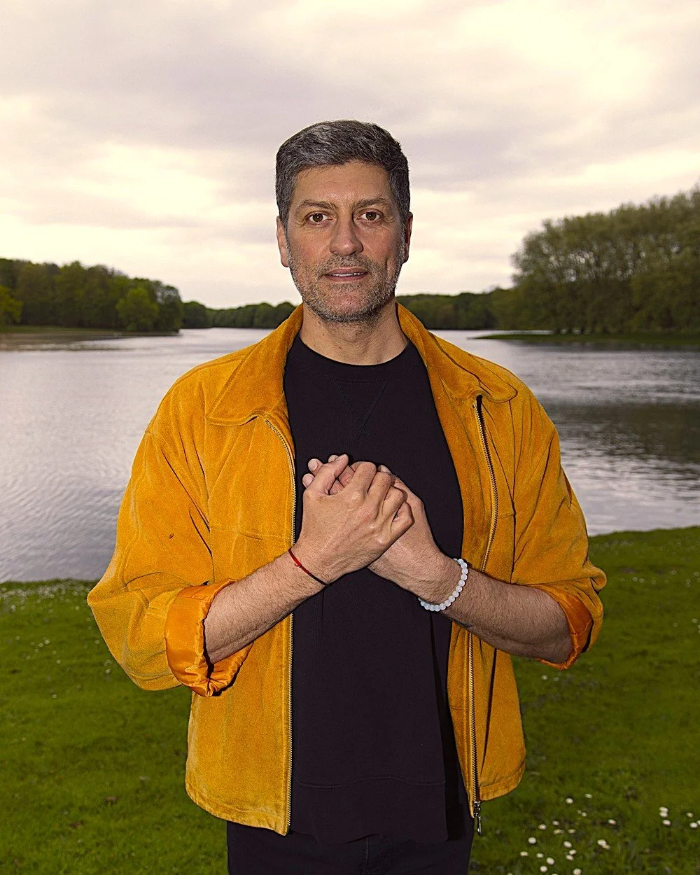 A man with gray hair and a beard, wearing a black shirt and a yellow jacket, standing outdoors near a body of water with trees in the background, holding his hands over his chest.
