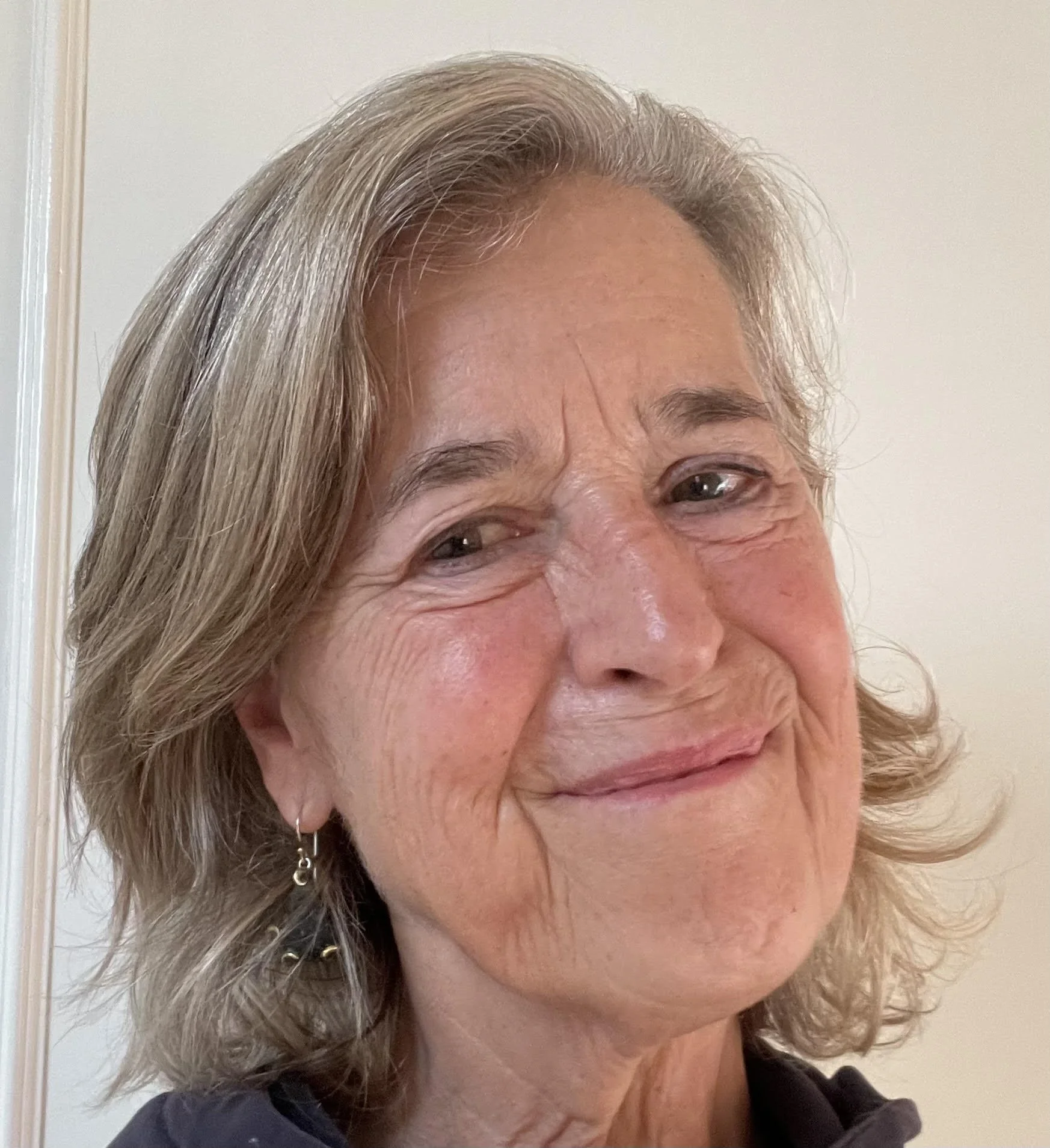 Close-up of an older woman with gray hair, smiling and looking at the camera, against a neutral background.