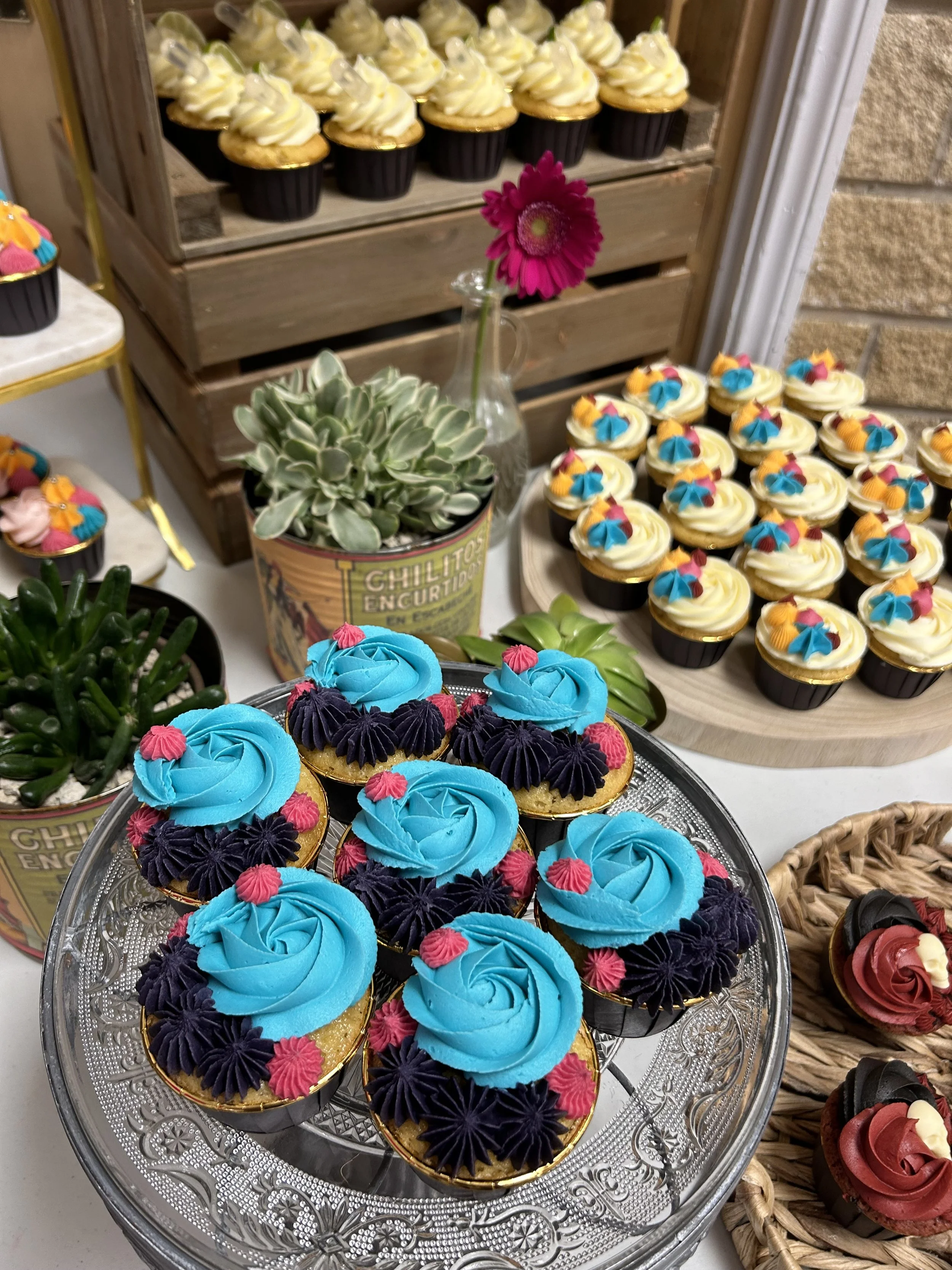 Display of cupcakes with blue and dark purple frosting decorated with small pink embellishments, arranged on a glass stand. Additional cupcakes with colorful frosting are visible in the background.