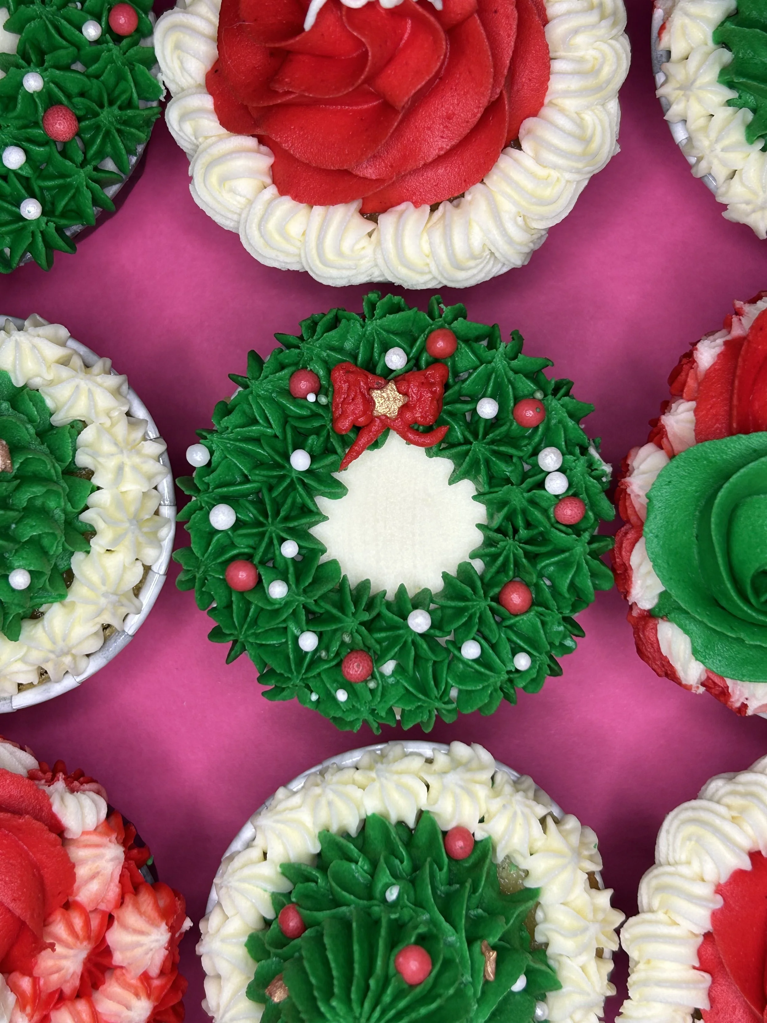 Decorative Christmas cupcakes with green, red, and white frosting, shaped like holly leaves, flowers, and wreaths, on a pink background.