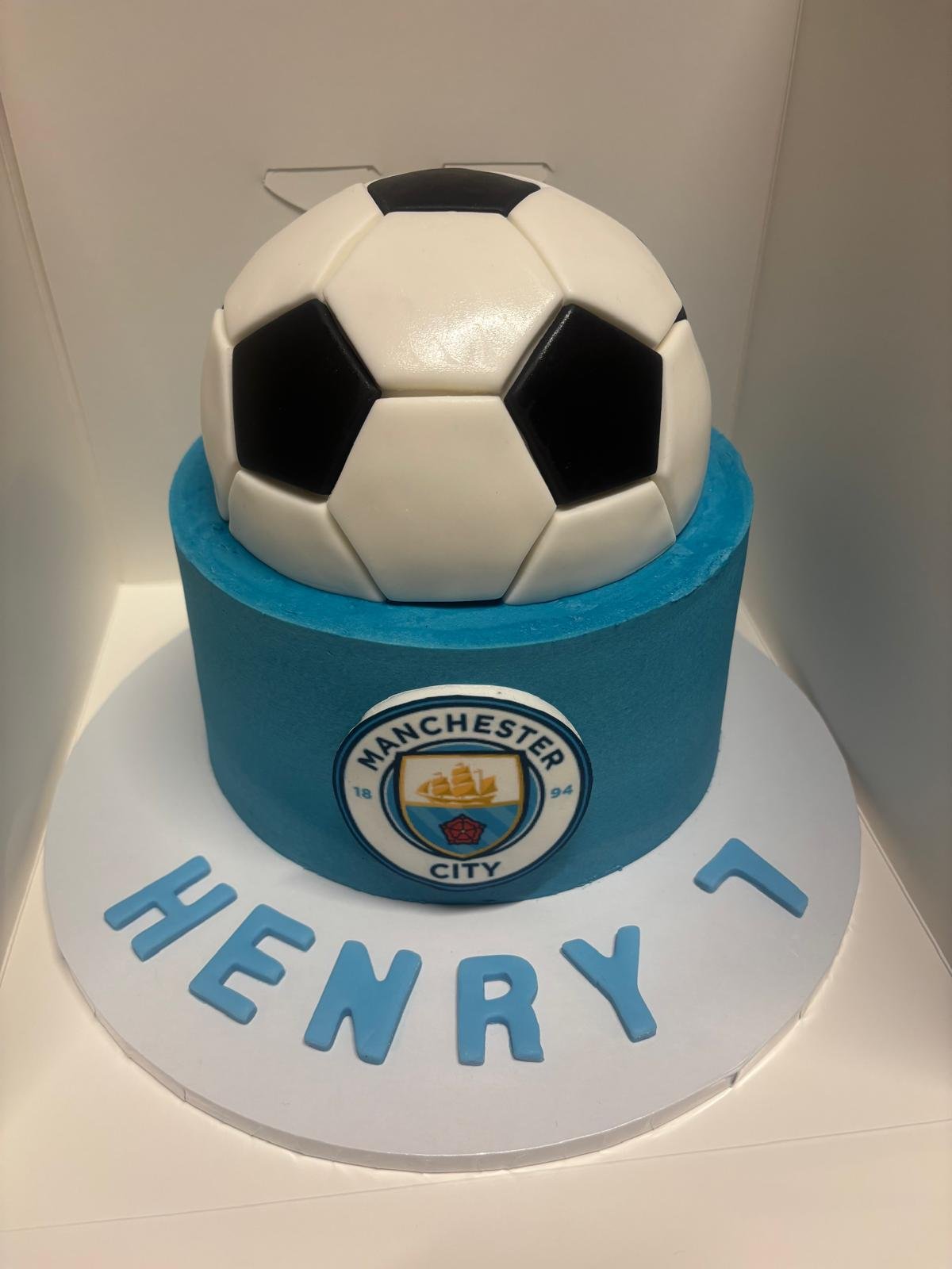 A birthday cake with a black and white soccer ball on top, decorated with the Manchester City Football Club logo, and the name 'HENRY' written in blue letters on a white base.