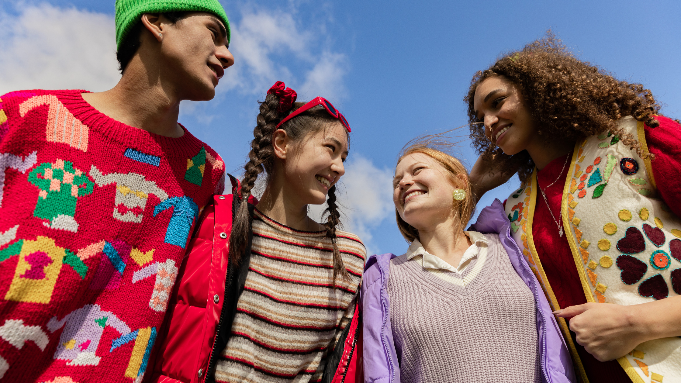 Four young people standing close together outdoors, smiling and laughing, with a blue sky behind them.