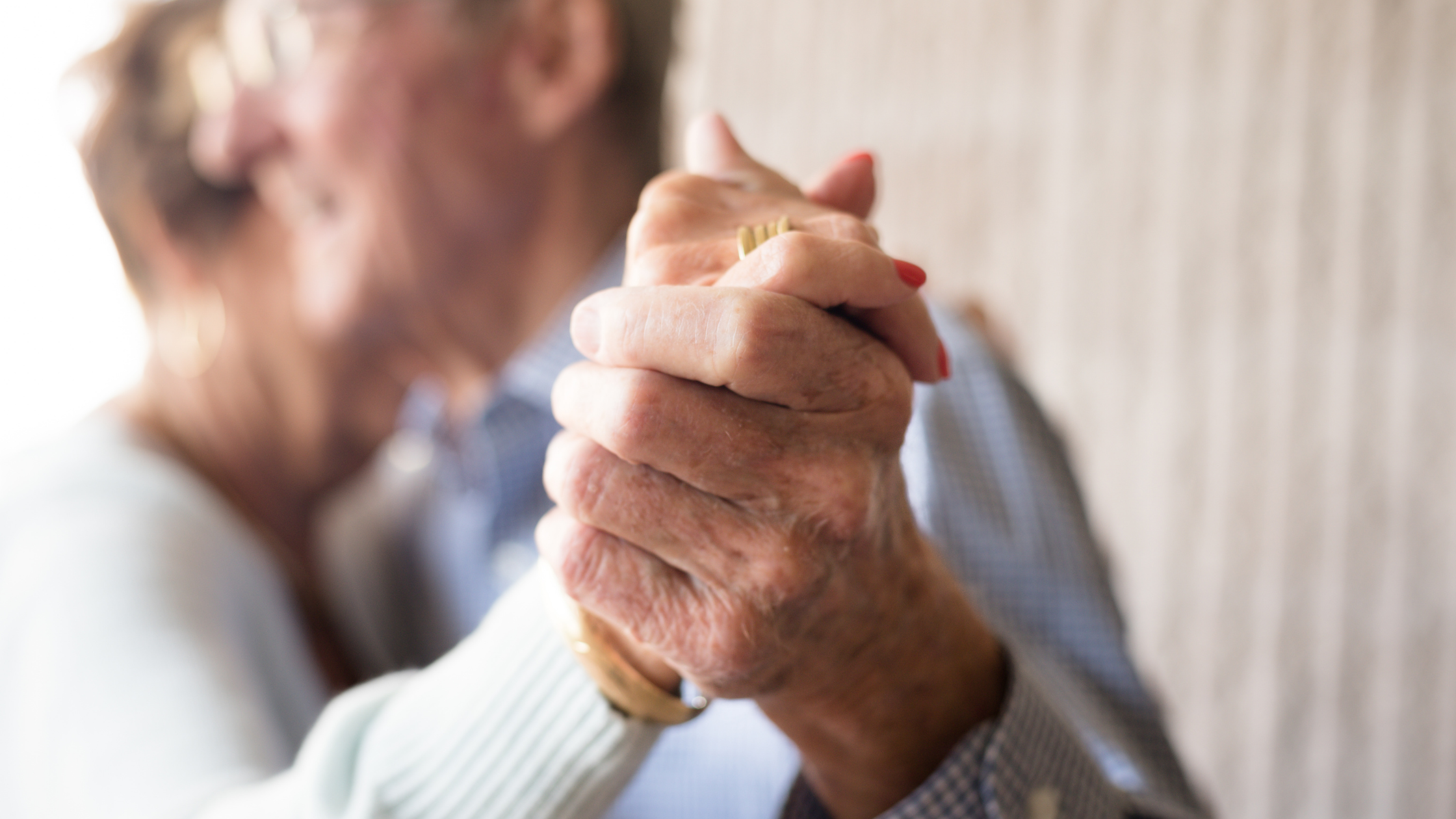A close-up of two pairs of hands clasped together, with two people visible in soft focus in the background.