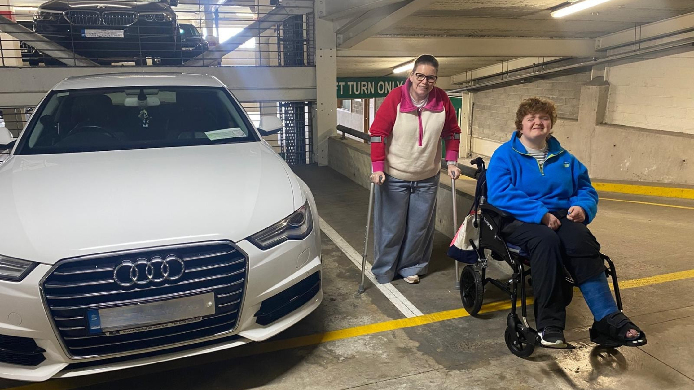 Two people in a multi-storey car park. Person standing wearing glasses and a burgundy and cream top. Person seated in a wheelchair wearing a blue jacket. White Audi car visible on the left.