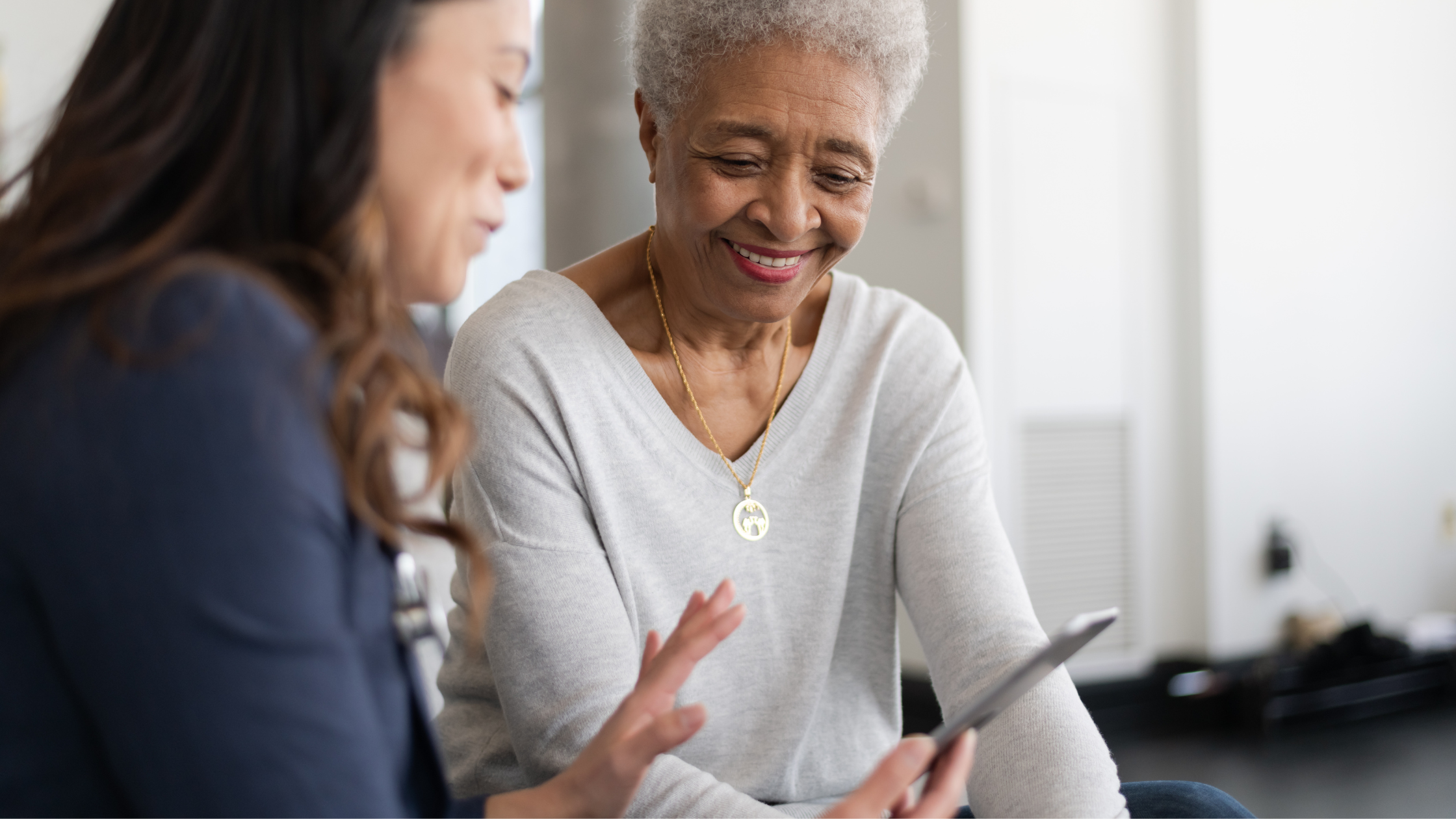 A person with short grey hair smiles warmly while looking at a tablet, seated next to another person who is gesturing towards the screen.