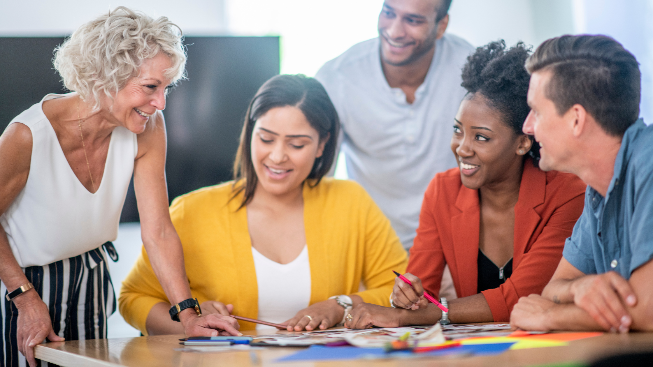 Five people gathered around a table in an office, smiling and looking at documents and materials spread across it.