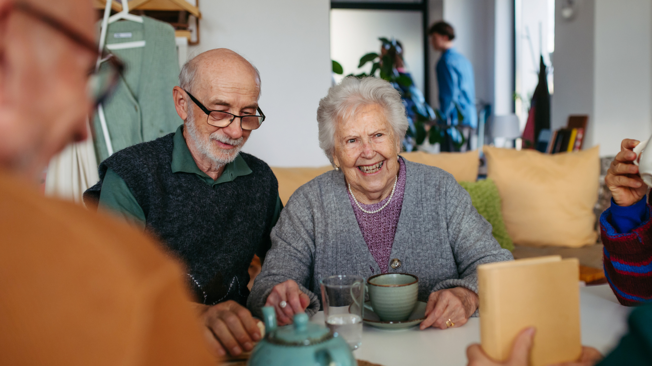 Two people sitting at a table with cups of tea, laughing together, with others visible in the background.