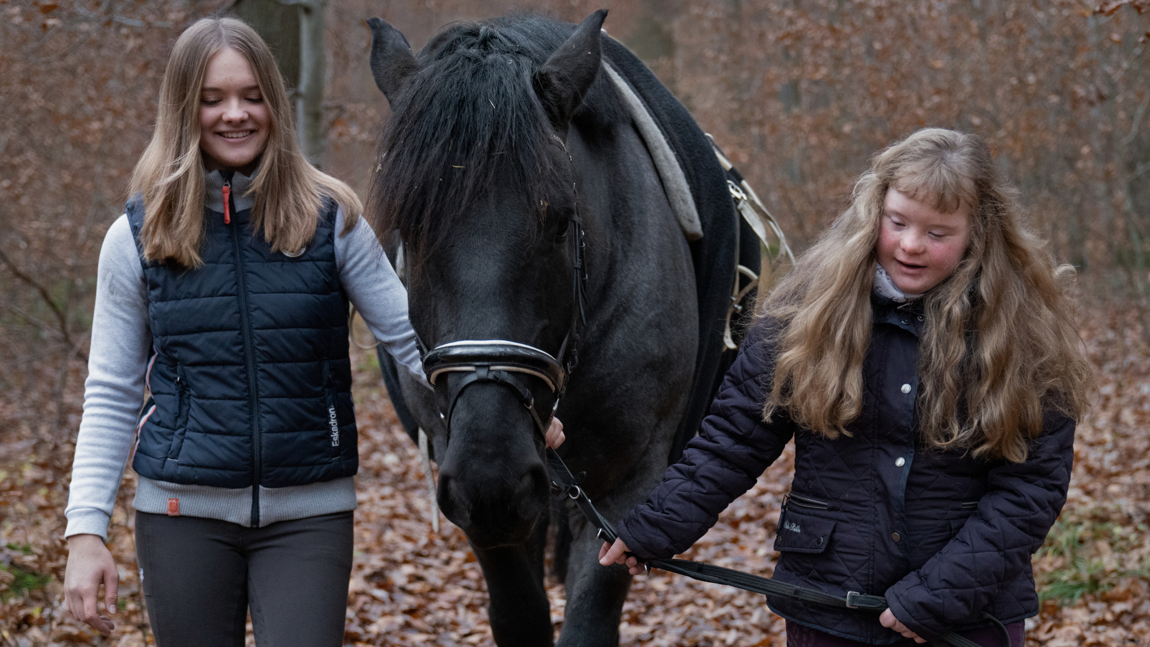 Two young people with black pony outdoors in woodland setting with autumn leaves on ground, both wearing dark jackets.