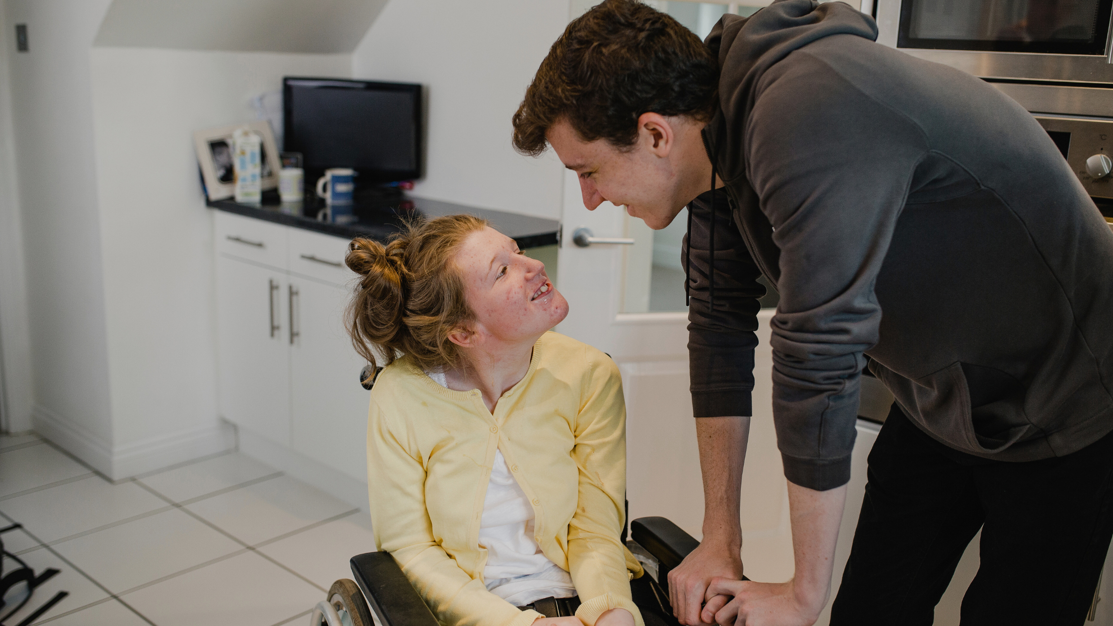 Wheelchair user in yellow cardigan and person in grey hoodie sharing joyful moment together in modern white kitchen.