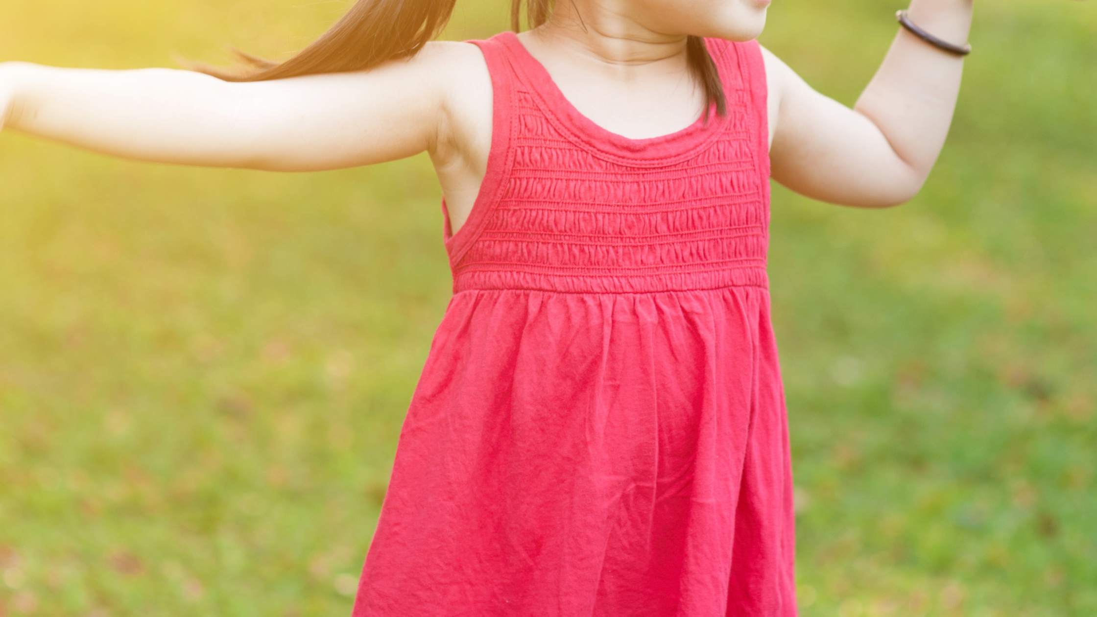 A child with arms outstretched outdoors on grass, face not visible, wearing a red dress.