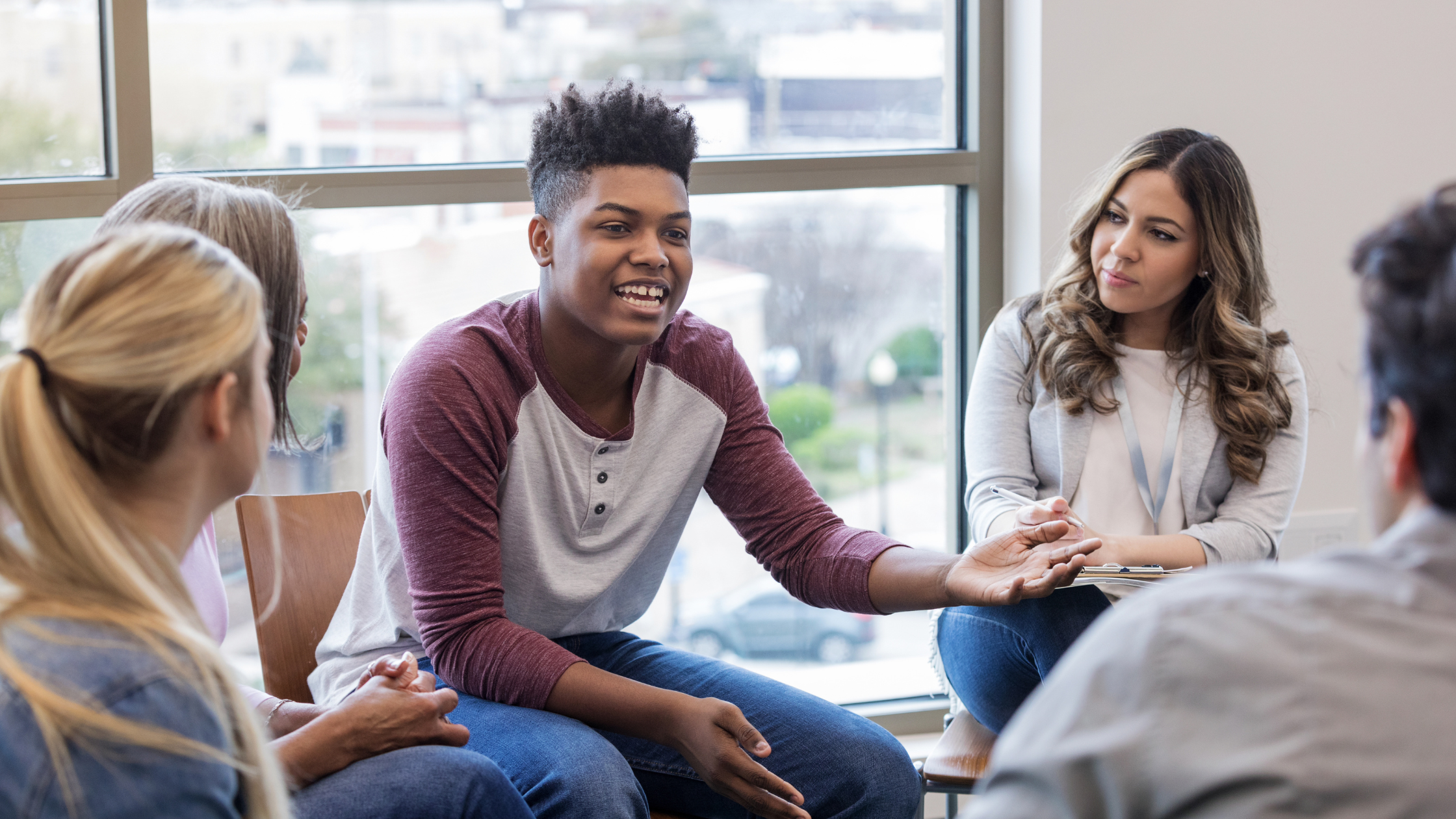 A group of people seated in a circle in conversation, one person speaking and gesturing expressively towards another.