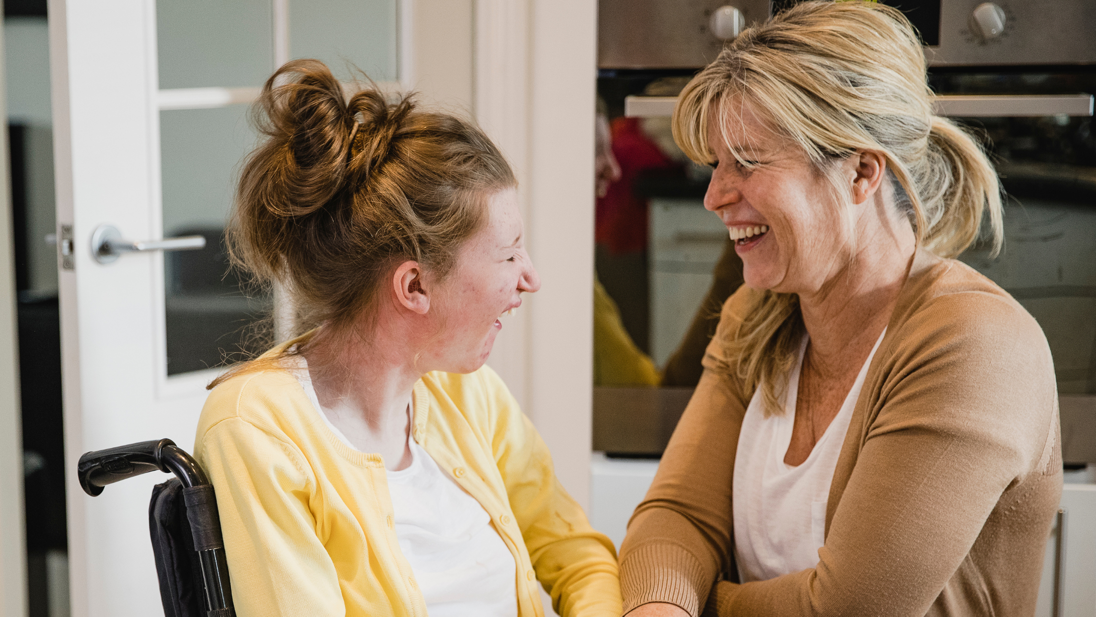 Wheelchair user in yellow cardigan and person in tan jumper laughing together in bright kitchen setting.
