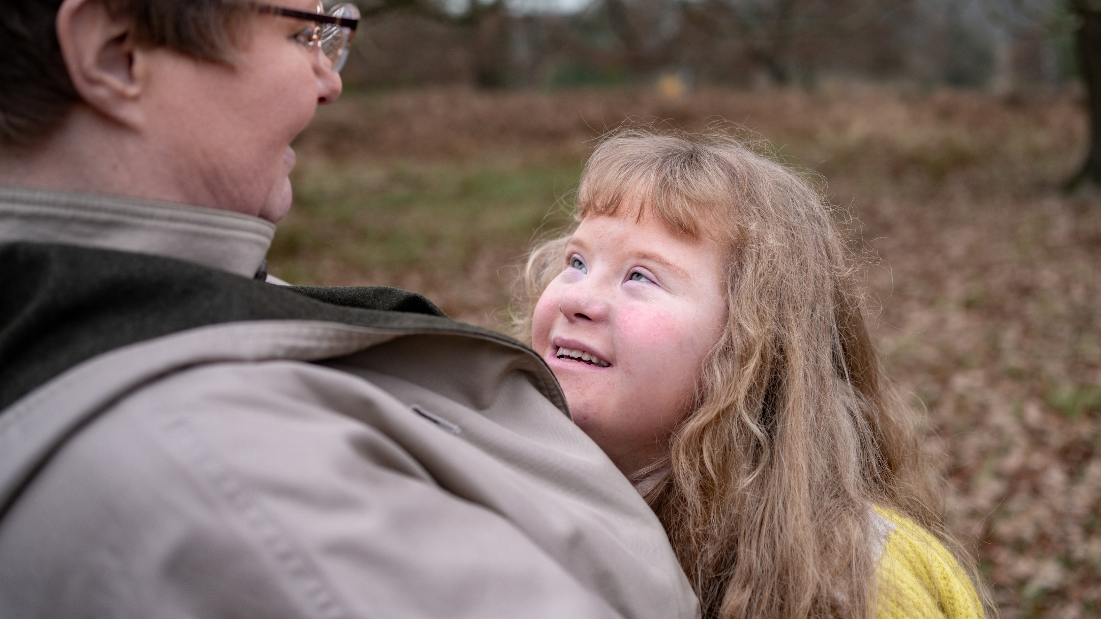 Child in yellow top smiling whilst looking up at person wearing glasses outdoors, autumn leaves on ground in background.