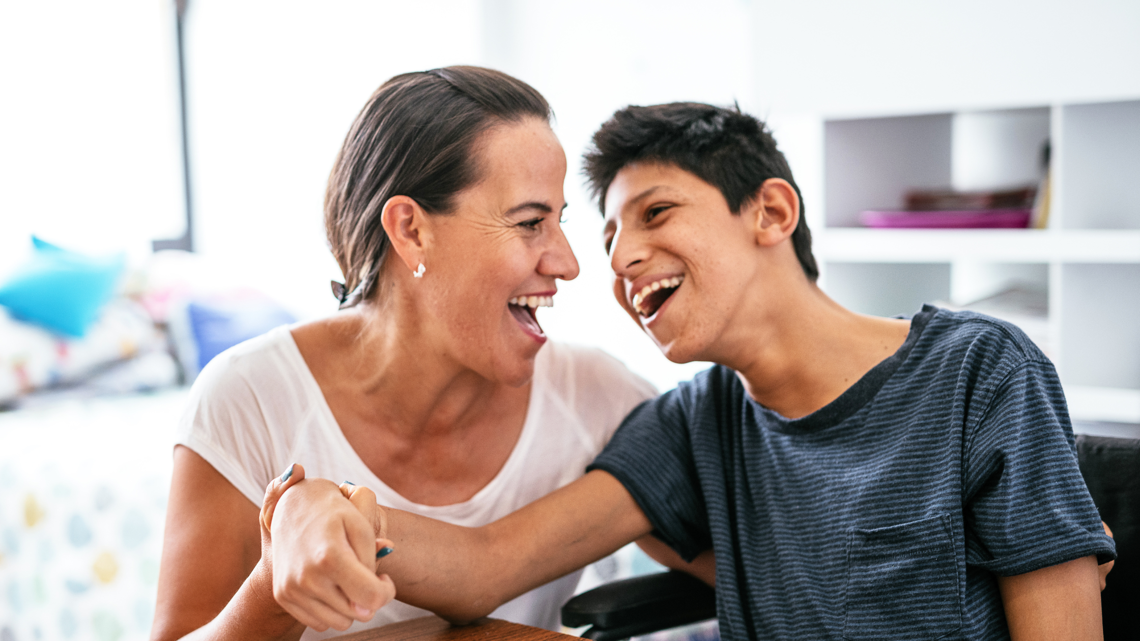 Two people laughing together indoors, one in white top, one in dark striped top, bright room with shelving in background.