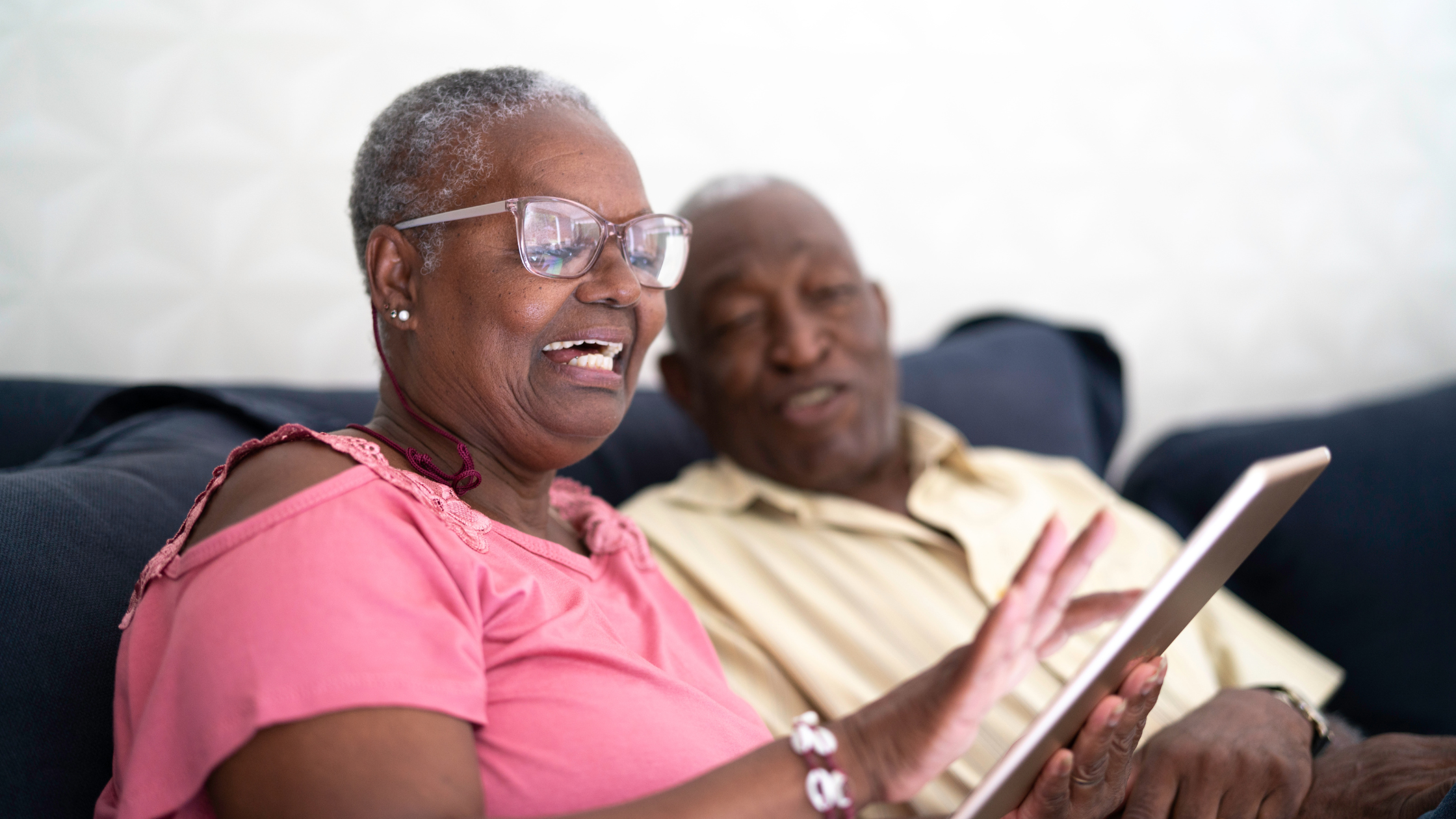 Two people on a sofa smiling whilst looking at a tablet together. Person in foreground wearing a pink top and glasses.