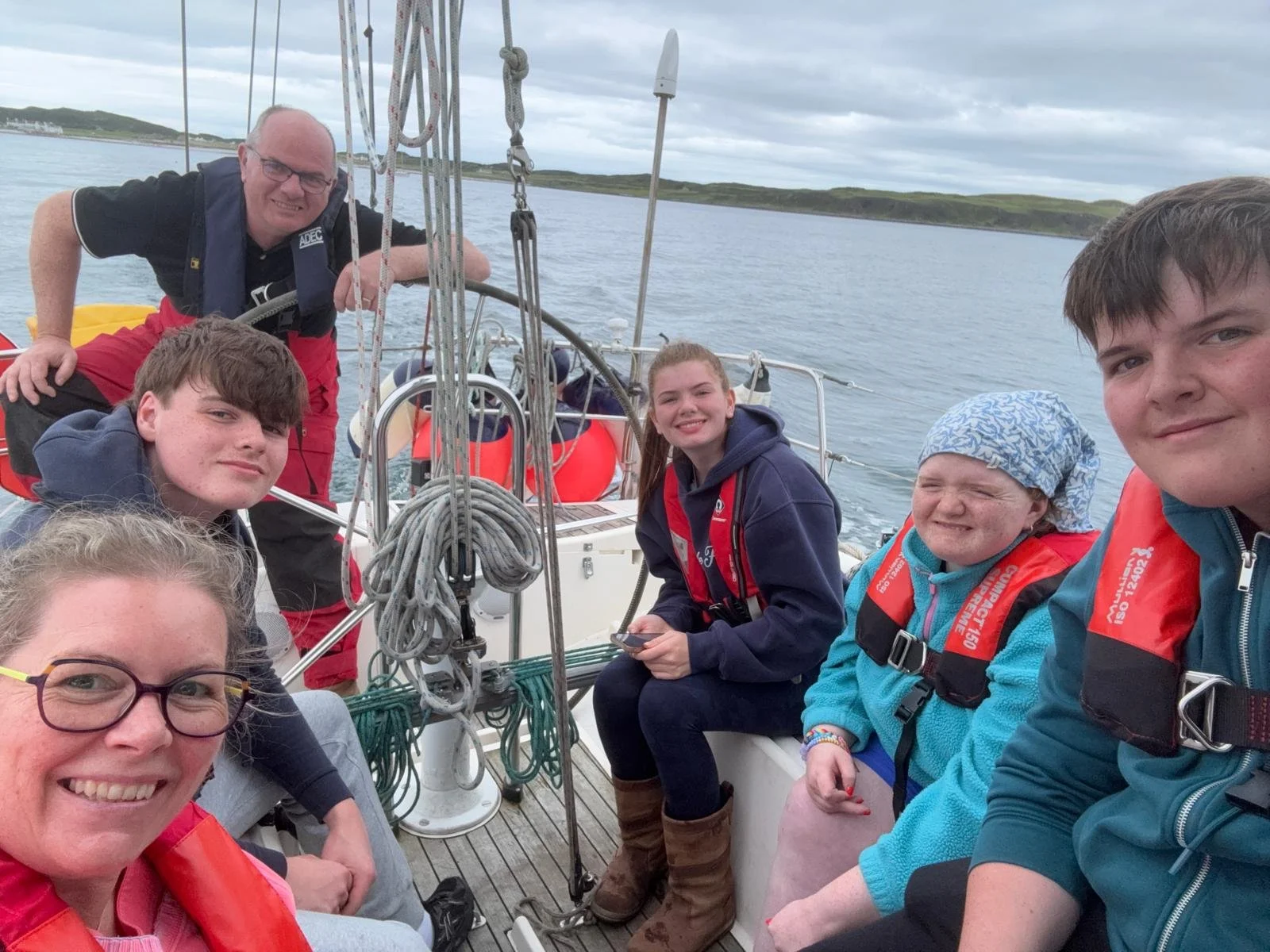 A family of five on the deck of a sailing boat, smiling at the camera, wearing life jackets, with open water and a distant coastline behind them.