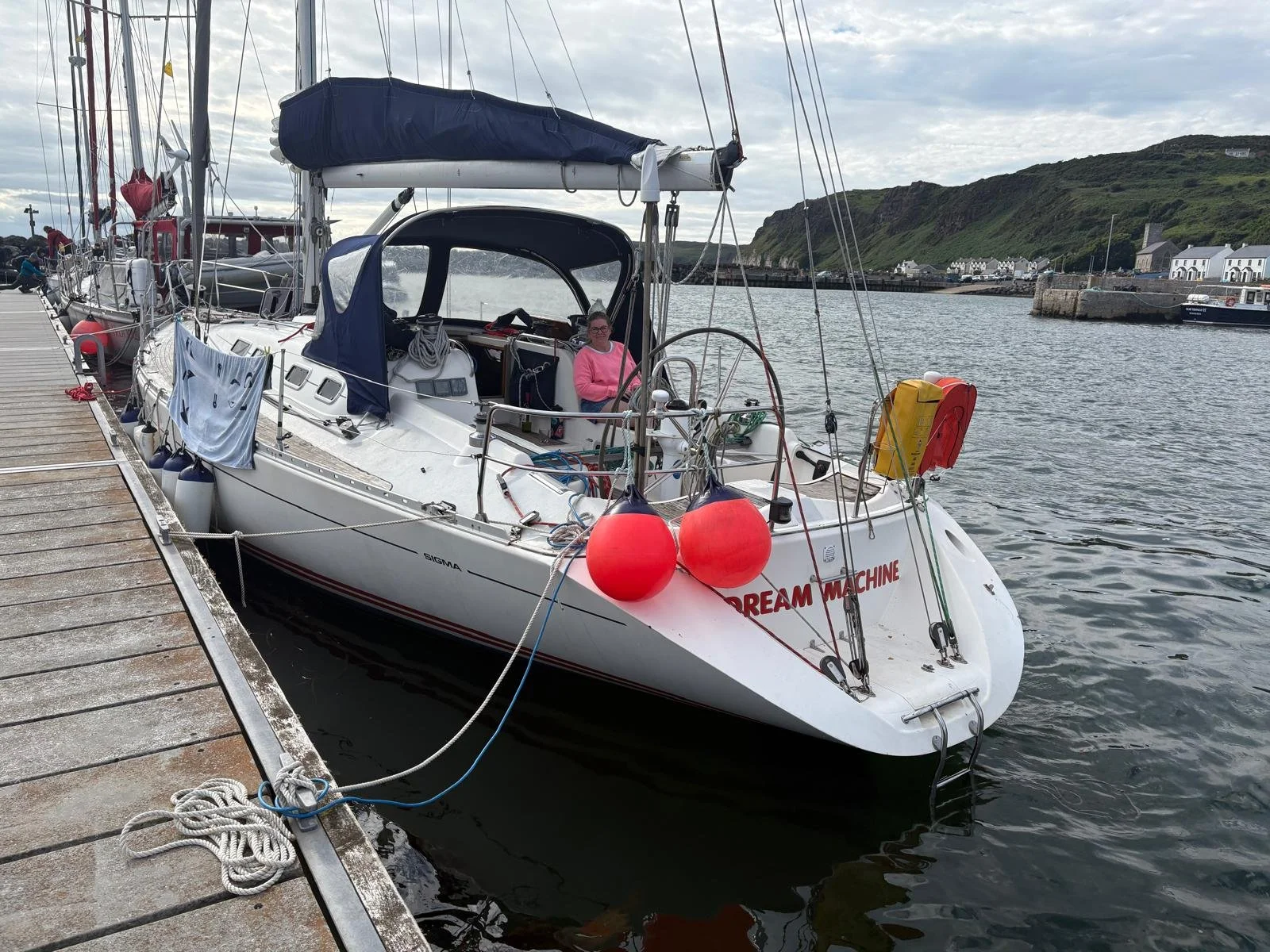 A white sailing boat named Dream Machine moored at a harbour pier, with green coastal cliffs and a small village visible in the background.