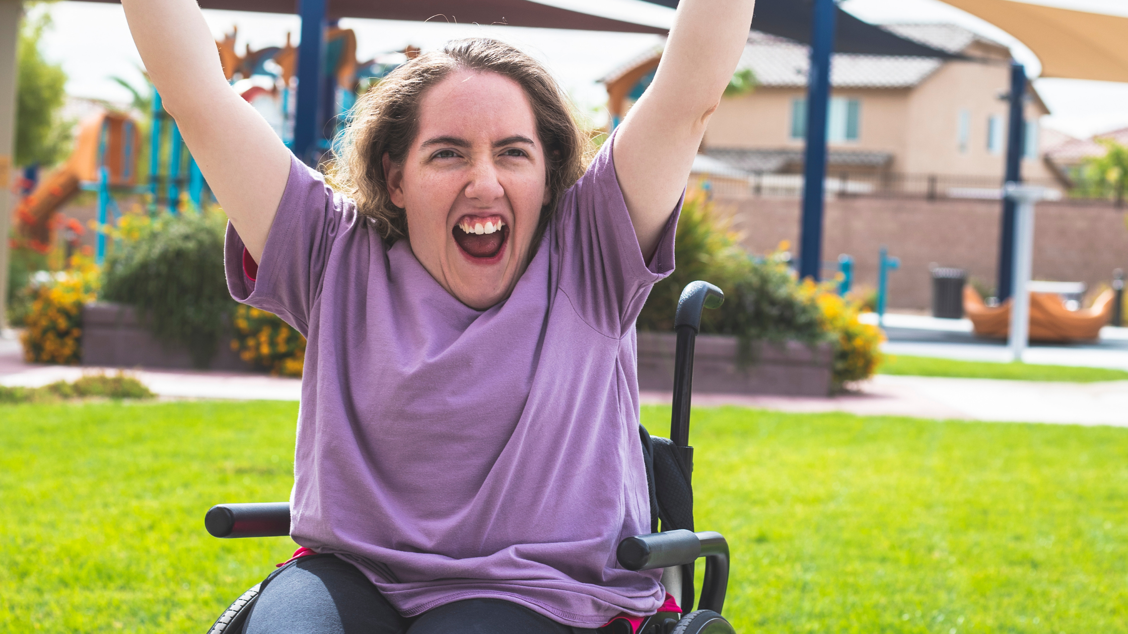 A person in a wheelchair outdoors, arms raised in celebration, laughing with a grassy park area visible in the background.