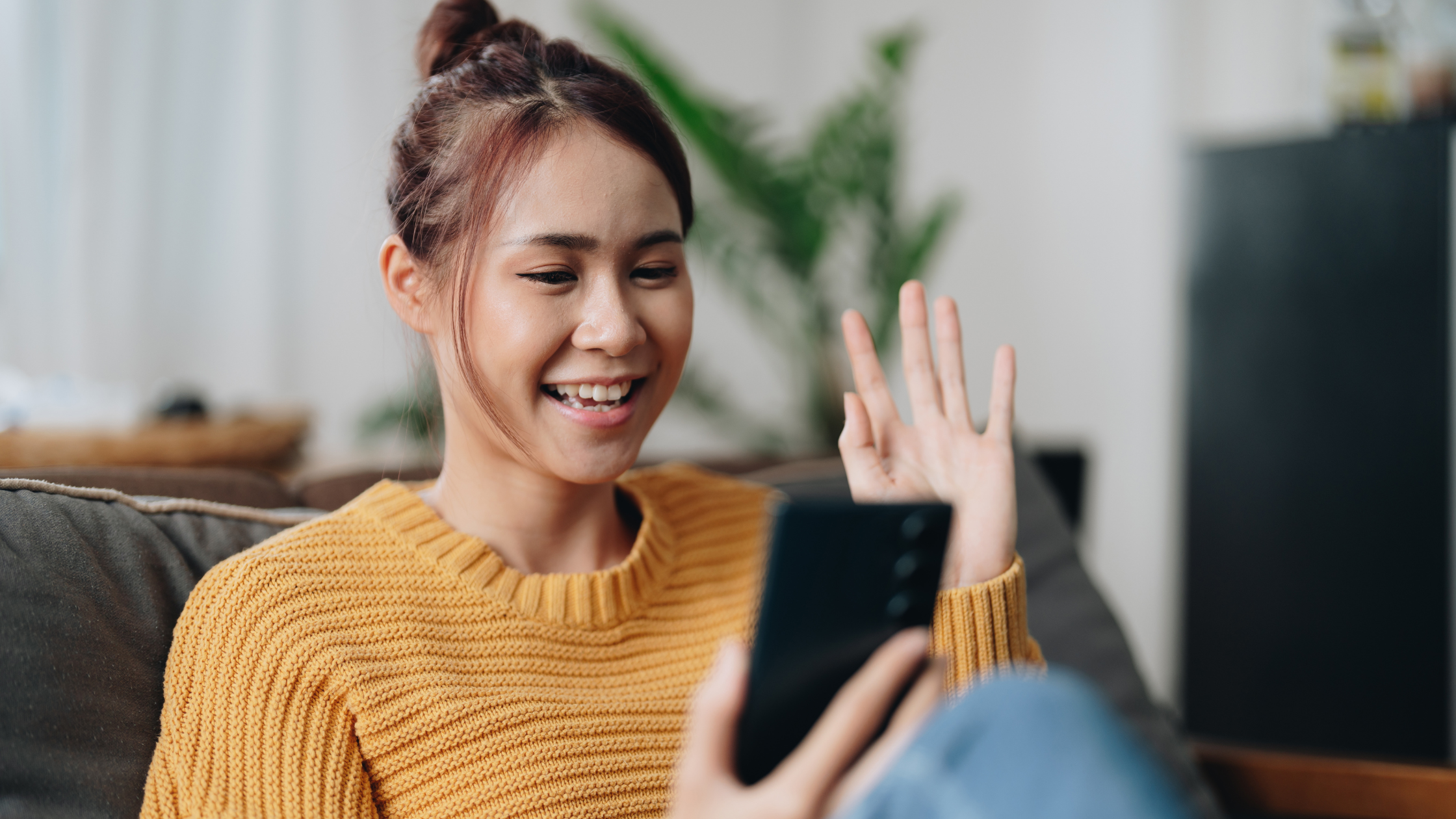 A person sitting on a sofa, smiling and waving at a smartphone during a video call.