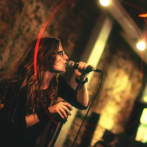 A woman with long dark hair and glasses singing into a microphone on stage during a performance.