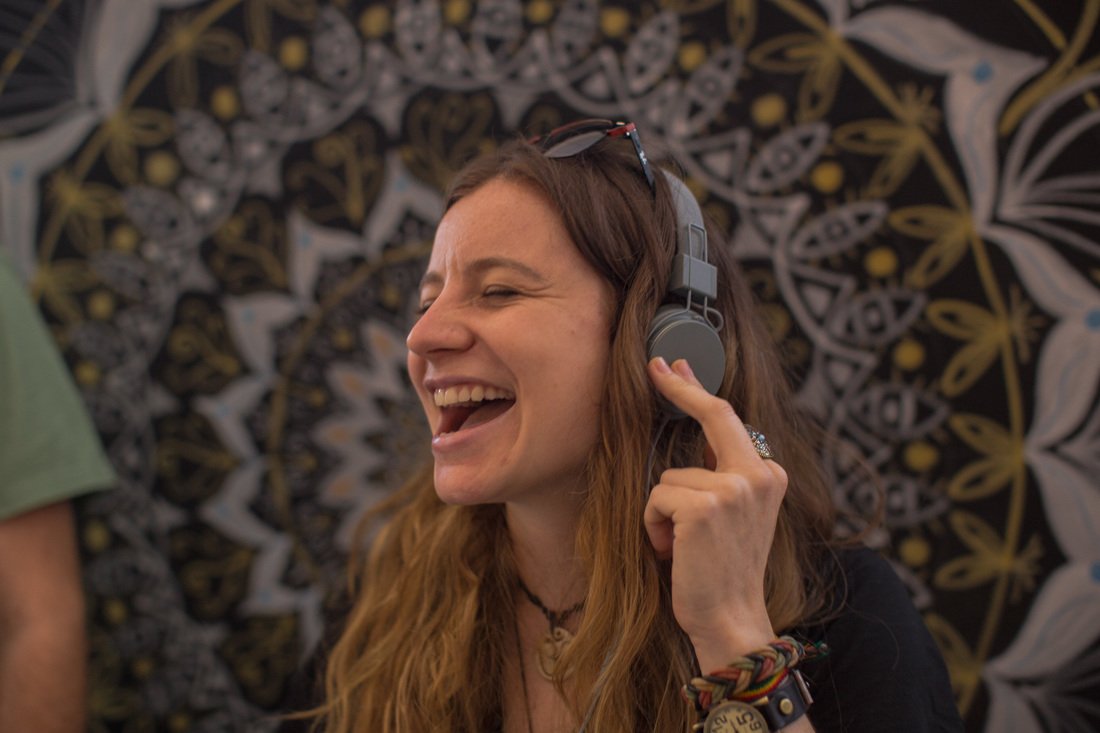 A woman with long, wavy hair wearing headphones and a black shirt is smiling and singing or speaking, with a patterned background behind her.