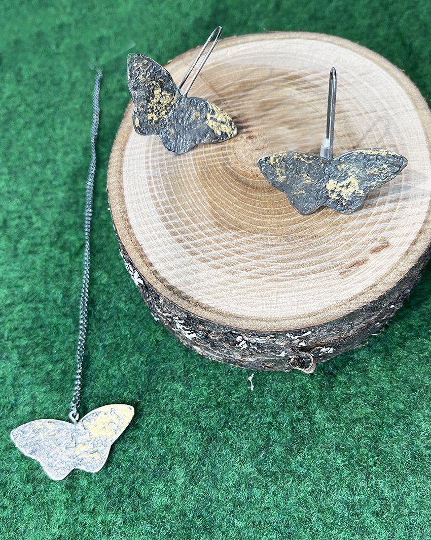 Små fjärilssmycken i metall på ett trästycke med grönt tyg i bakgrunden.