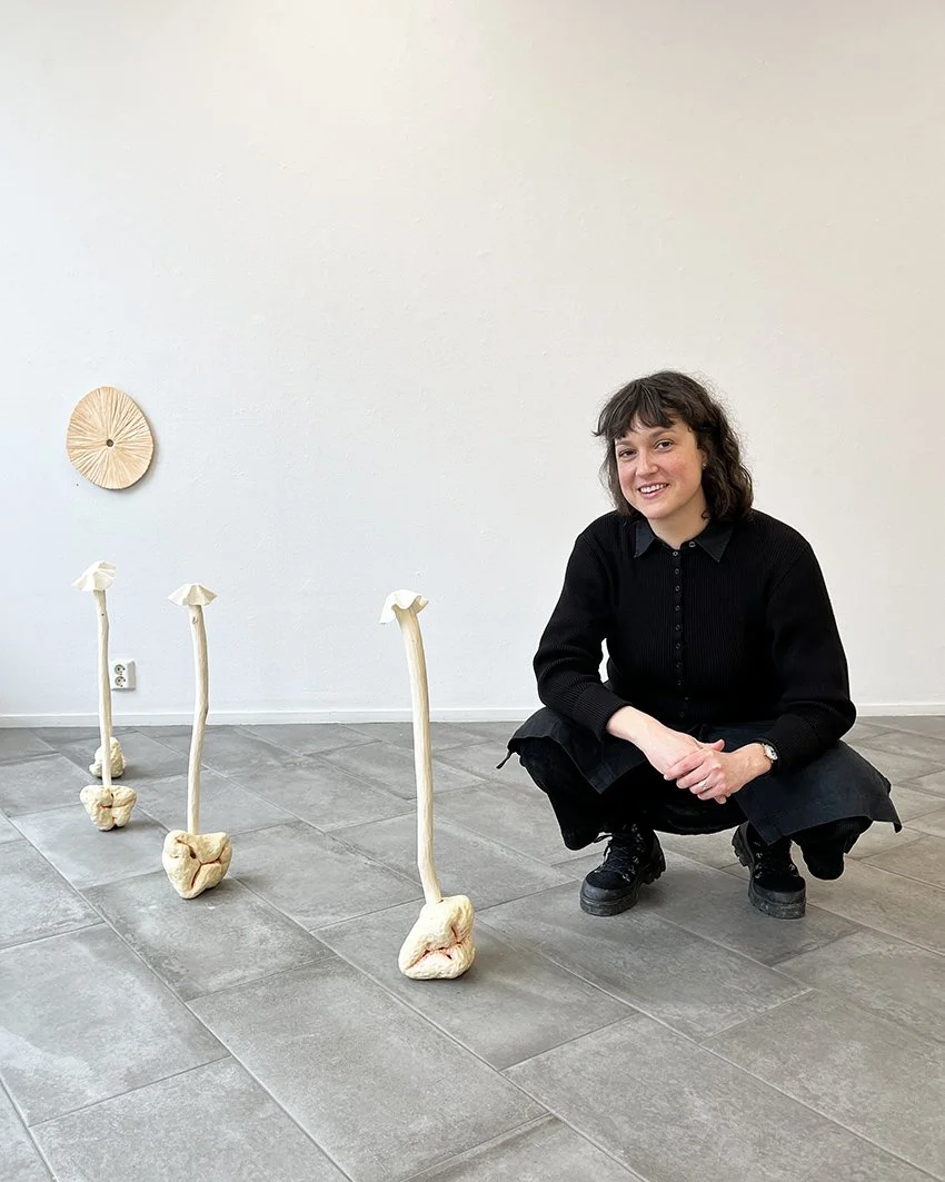 En kvinna i svart outfit som är knäböjd bredvid tre konstgjorda ben med sgraffitiga fötter på en stengolv i ett konstgalleri.